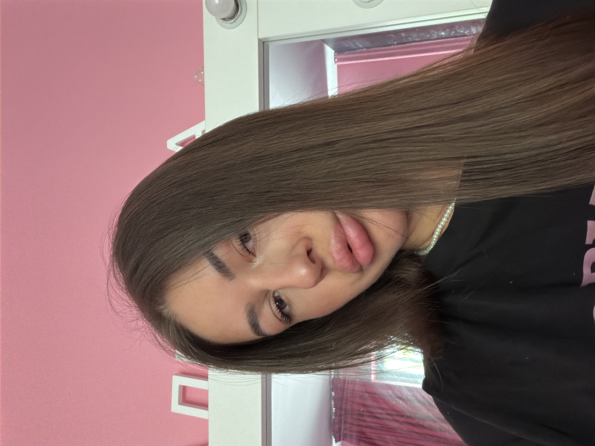 A young woman is captured in a close-up, selfie-style photograph, facing the camera with her head slightly tilted. She has long, straight, dark brown hair that frames her face. Her lips are noticeably full and slightly parted, and she appears to be wearing a string of pearls around her neck. She is dressed in a black t-shirt. The background consists of a pink wall with white decorative letters, and a white frame that suggests a mirror or a lighted vanity. The overall environment appears to be an indoor, likely personal space, such as a bedroom or dressing area, with hints of a pink curtain visible in the reflection of the frame. No specific geographical markers definitively place this image in Novi Iskar, Bulgaria.