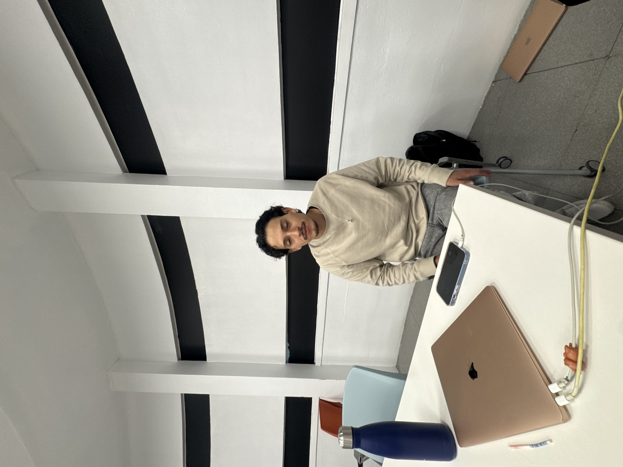 A male is sitting at a table with a laptop, phone, and water bottle in what appears to be a modern, brightly lit room. The room has distinctive architectural features including white walls with black stripes on the ceiling. The location is not specified as Puebla, Mexico, nor does the image provide data to suggest this.