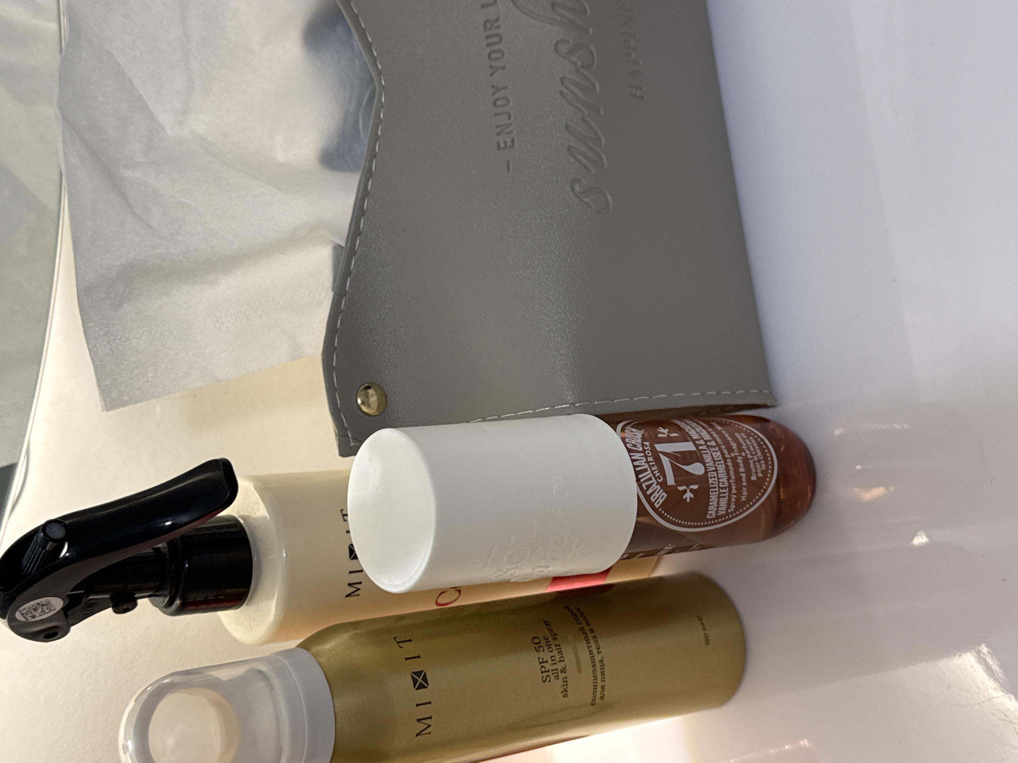 The scene depicts three cosmetic bottles alongside a grey tissue box on a white surface. The bottle closest to the foreground is a golden-colored "MIXIT" brand SPF 50 all-in-one skin and hair spray. Above it is a white bottle with a black spray nozzle. The third bottle is brown and labeled "BRAZILIAN CRUSH CHEIROSA '71'" with "CARAMELIZED VANILLA & MACADAMIA" noted. The tissue box is partially visible and has the text "- ENJOY YOUR L sunsh HAPPINE". The bottles appear to be resting on a white surface. The location and city Bugry, Russia are not evident in the image.