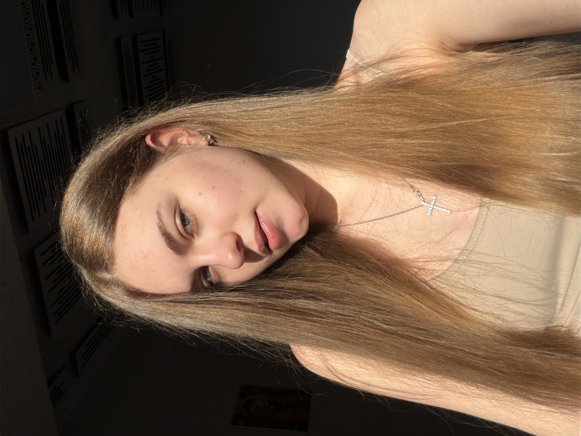 The media file is a close-up, angled selfie of a young woman with long, light brown hair. She is wearing a light-colored camisole and a silver necklace with a cross pendant. The lighting suggests it is daytime, with strong sunlight casting shadows. The background is dark, with some wall decorations visible on the left, appearing to be acoustic panels. The overall mood seems relaxed and natural. No text is visible.