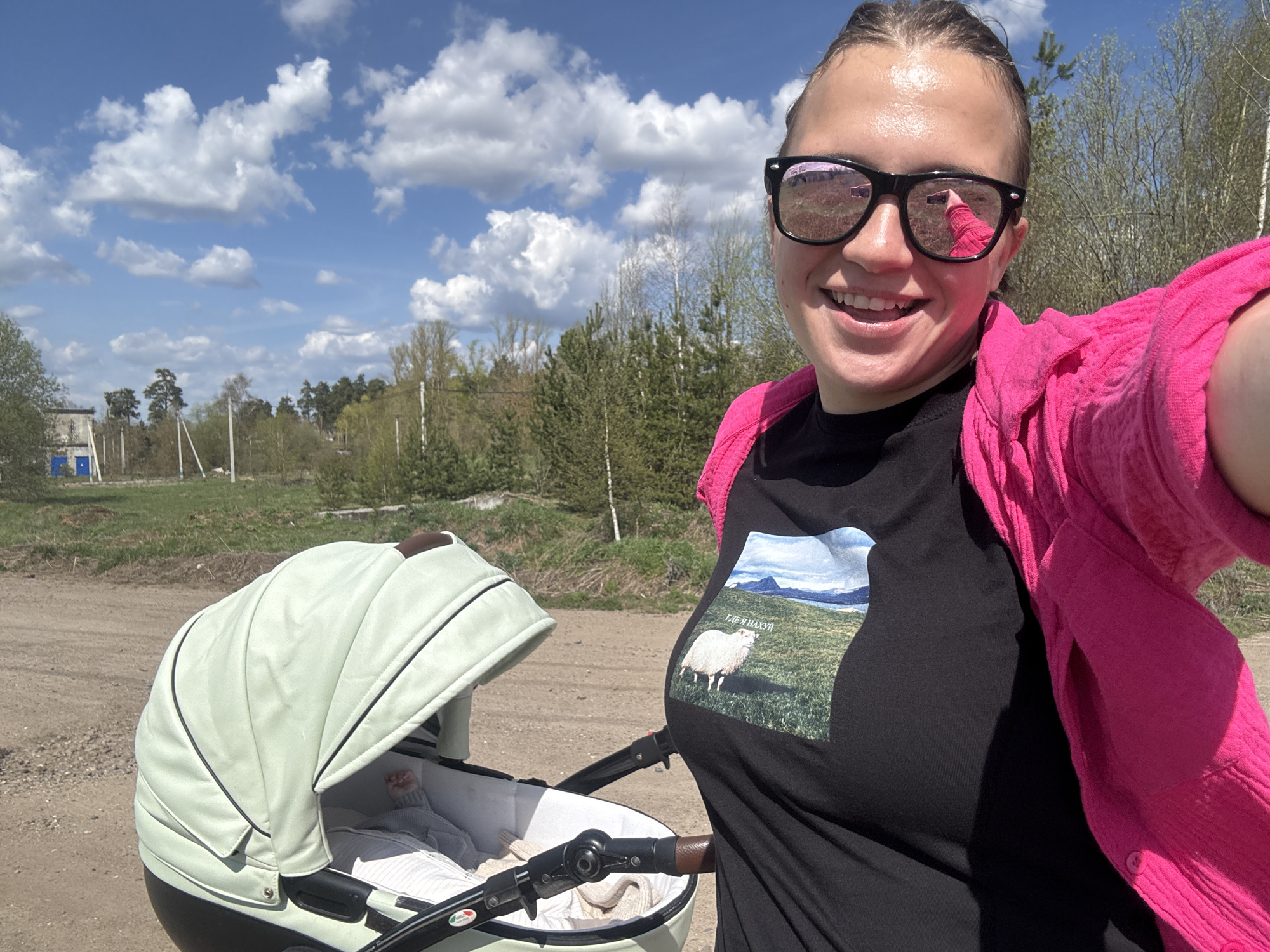 A woman is taking a selfie outdoors, likely on a dirt road in Раздолье, Russia, based on the location tag. She is wearing sunglasses, a black t-shirt with an image of a sheep and landscape, and a pink cardigan. Beside her is a light green baby stroller with a baby inside. The background features a field, trees, blue sky with clouds, and some buildings in the distance.