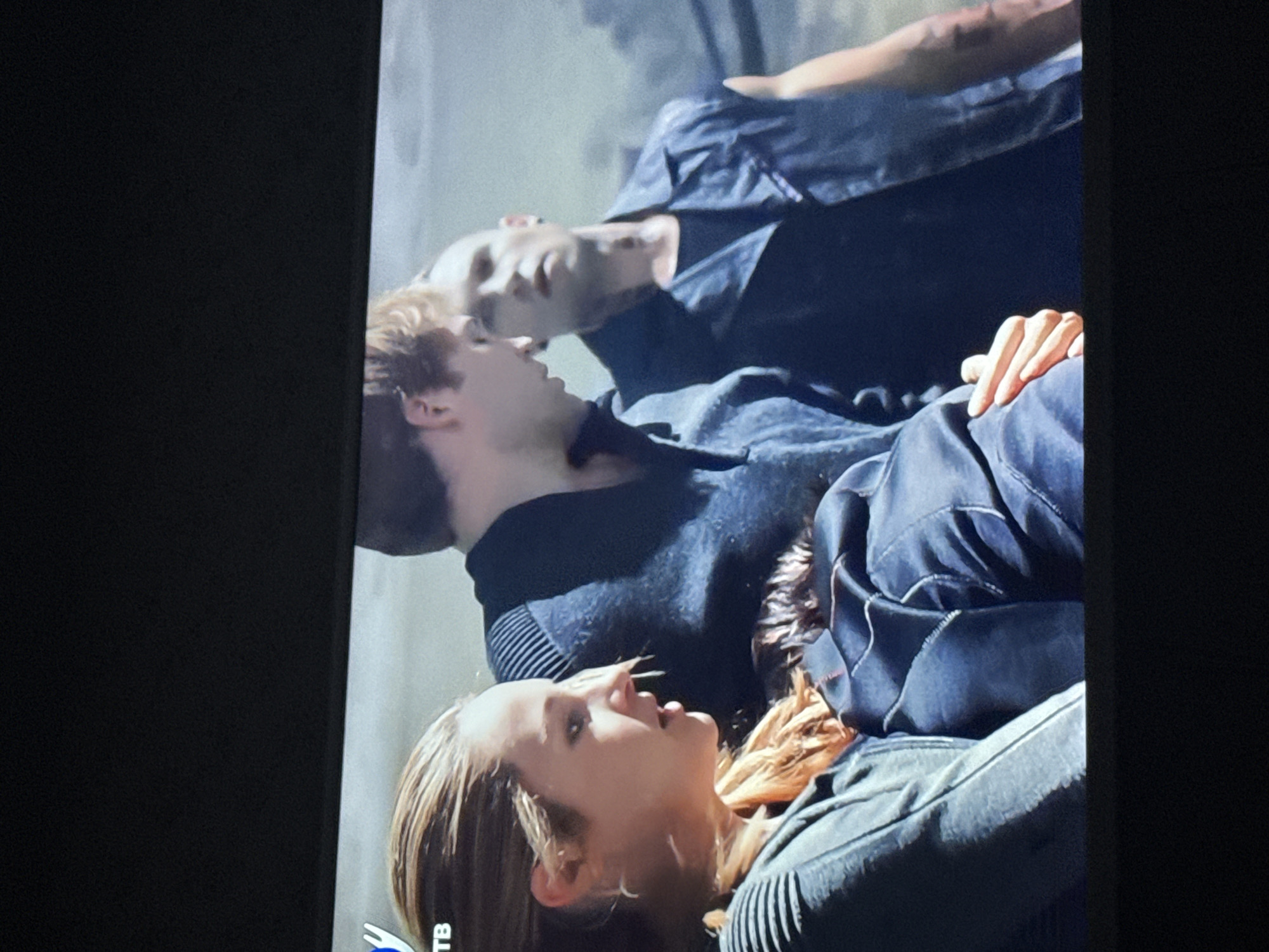 Three people are lying down. The person closest to the camera is a fair-skinned woman with blonde hair, wearing a dark-colored garment with ribbed detailing. Behind her is a fair-skinned man with short reddish-brown hair, also wearing a dark-colored garment. Behind him is a dark-skinned man, mostly bald, wearing a dark-colored garment, his arm visible. The location and city are unknown.