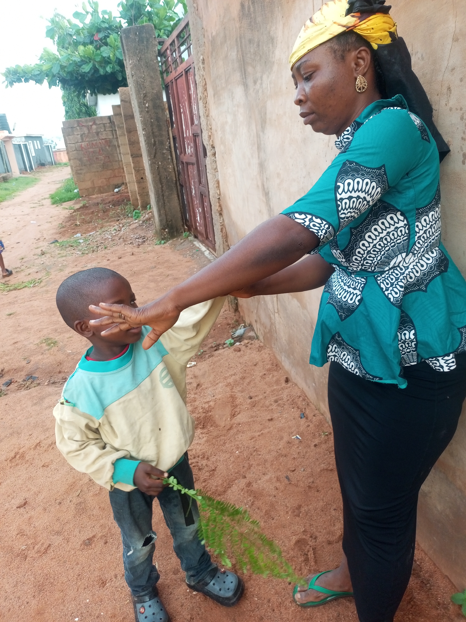 In Tempure, Nigeria, a woman wearing a yellow headscarf and a green and white patterned tunic stands leaning against a tan wall. She has her right arm extended, with her hand touching the face of a young boy. The boy, wearing a beige and light blue long-sleeved shirt and ripped blue jeans, is shielding his eyes with his left hand and holding a green leafy branch. The ground is covered in red dirt, with some scattered debris and a dusty path leading into the background. A rusty metal gate is visible on the left side of the wall, and trees with green foliage are in the distance.