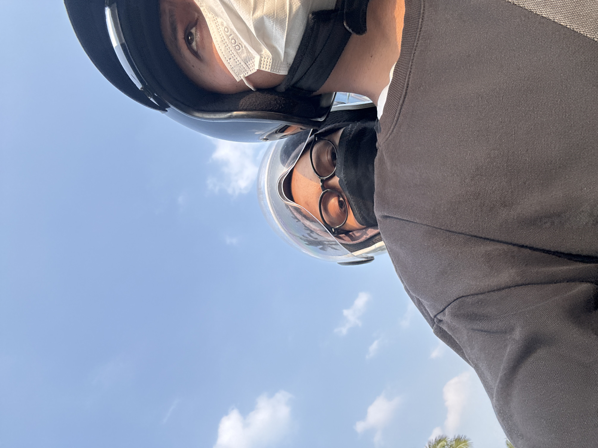 Two individuals wearing helmets and face masks are riding a motorcycle against a blue sky with scattered clouds, captured in a selfie-style shot. The person in the foreground, with a black helmet and a white mask, is looking upwards. Behind them, a second person with glasses and a black mask is visible. The scene appears to be in Pakulonan Barat, Indonesia, as indicated by the context, with a hint of trees visible on the left side.