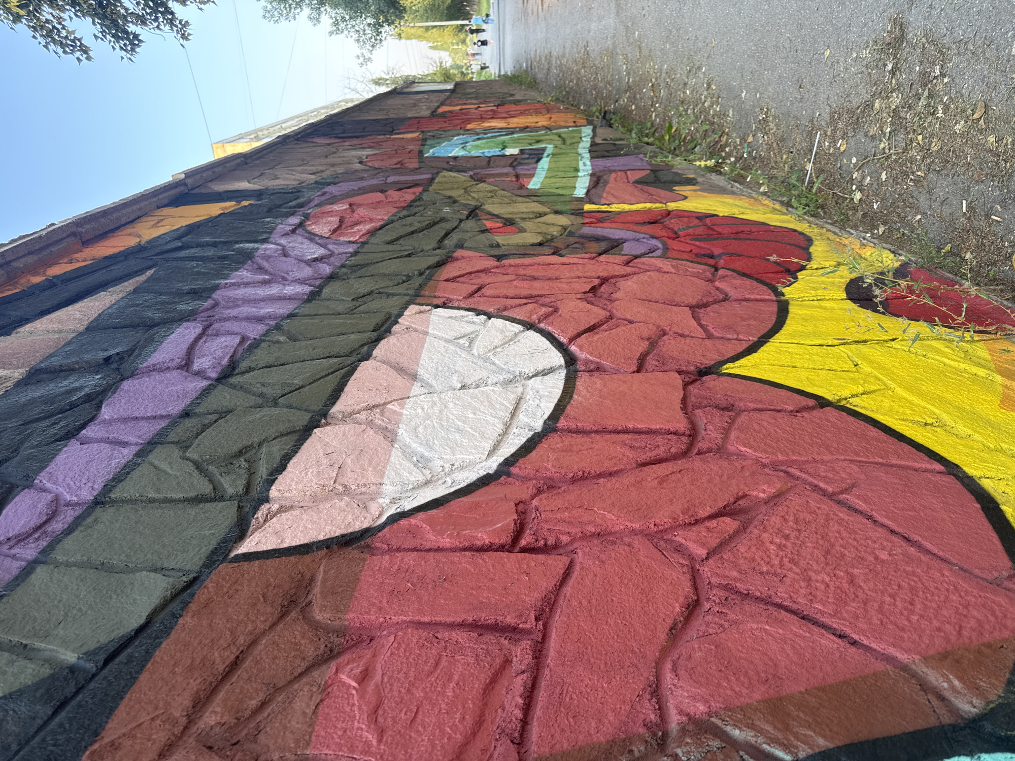 The image shows a section of a vibrantly painted stone wall, likely part of an underpass or retaining wall, in Ufa, Russia. The wall is adorned with a colorful graffiti mural featuring geometric patterns and abstract shapes in hues of red, yellow, purple, black, green, and white. The textured stone surface of the wall is clearly visible beneath the paint.
To the right of the wall is a paved pathway, possibly a pedestrian walkway or a service road. The pathway is lined with sparse vegetation and fallen leaves. In the background, several people are walking along the pathway, some appearing to be adults and possibly children. The sky is clear and blue, indicating daytime. A tree with foliage is visible in the upper right corner of the frame. The overall scene depicts an urban environment with public art integrated into the infrastructure.