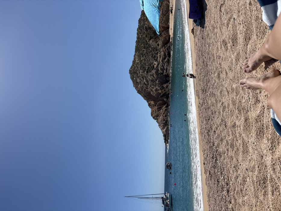 A sunny beach scene with a large rocky outcrop on one side and clear blue water meeting the sandy shore. Several boats and people are in the water and on the beach. A pair of feet is visible in the lower right corner on a towel. The clear blue sky takes up a significant portion of the image.