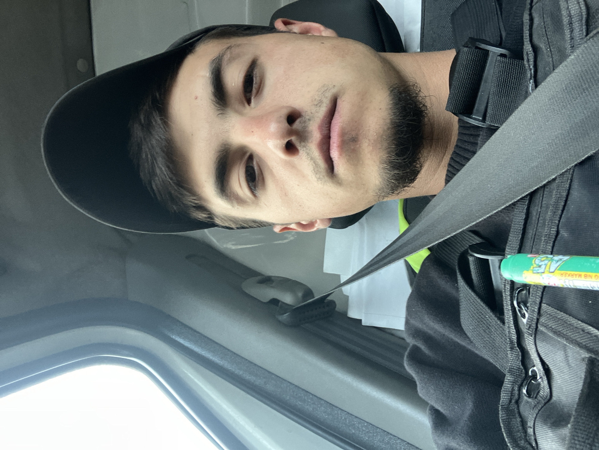 The media file displays a close-up, portrait-oriented shot of a young man, likely in his late teens or twenties, seated inside a vehicle. He has short dark hair, dark eyes, and a neat goatee and stubble on his face. He is wearing a dark baseball cap, a dark sweater or jacket, and a gray seatbelt diagonally across his chest. His expression is neutral and calm as he looks directly into the camera.
To his right, a dark utility bag or vest with straps and metal rings is visible, to which a bright green marker pen is clipped. Partial text on the marker reads "G NIB MARKER" and "45". Behind him, a light-colored seat headrest and a stack of white papers or documents are discernible, suggesting a work-related context. The bright light entering from a window on the left side of the frame indicates it is daytime. The vehicle interior features gray and light-colored plastic panels, consistent with a car, van, or truck. The overall setting is inside a vehicle, possibly during a break from work or travel in Novo Selo, Bulgaria.