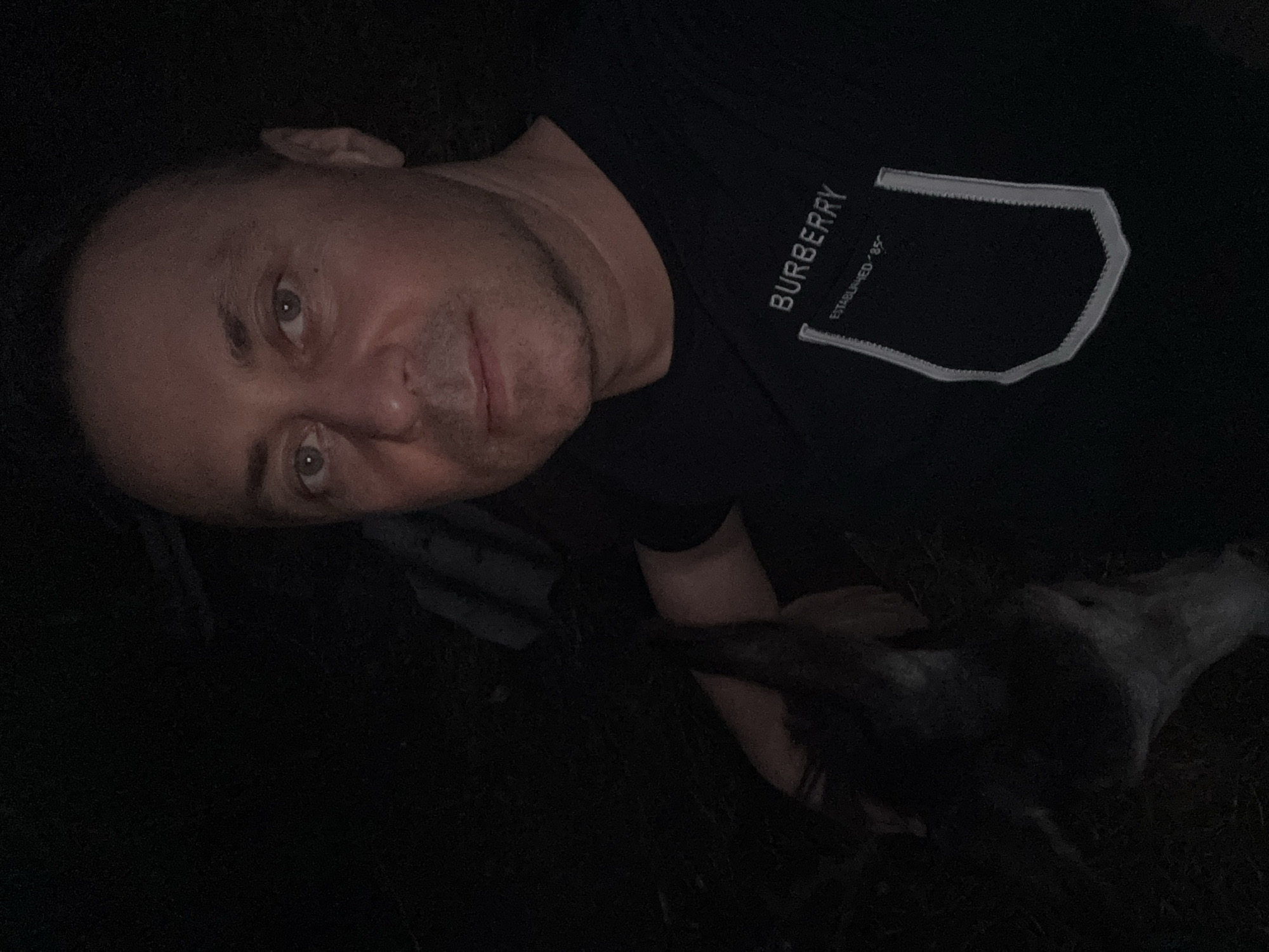 A selfie is taken by a man at night in an outdoor, dark environment. The man is wearing a black t-shirt with "BURBERRY" and "ESTABLISHED '85" printed on it. To the left of the man, a dark-colored animal's head is visible, appearing to be a dog. The background is mostly dark, suggesting it is night and possibly a rural or wooded area. There is no visible indication of the city of Tandzia, Georgia.