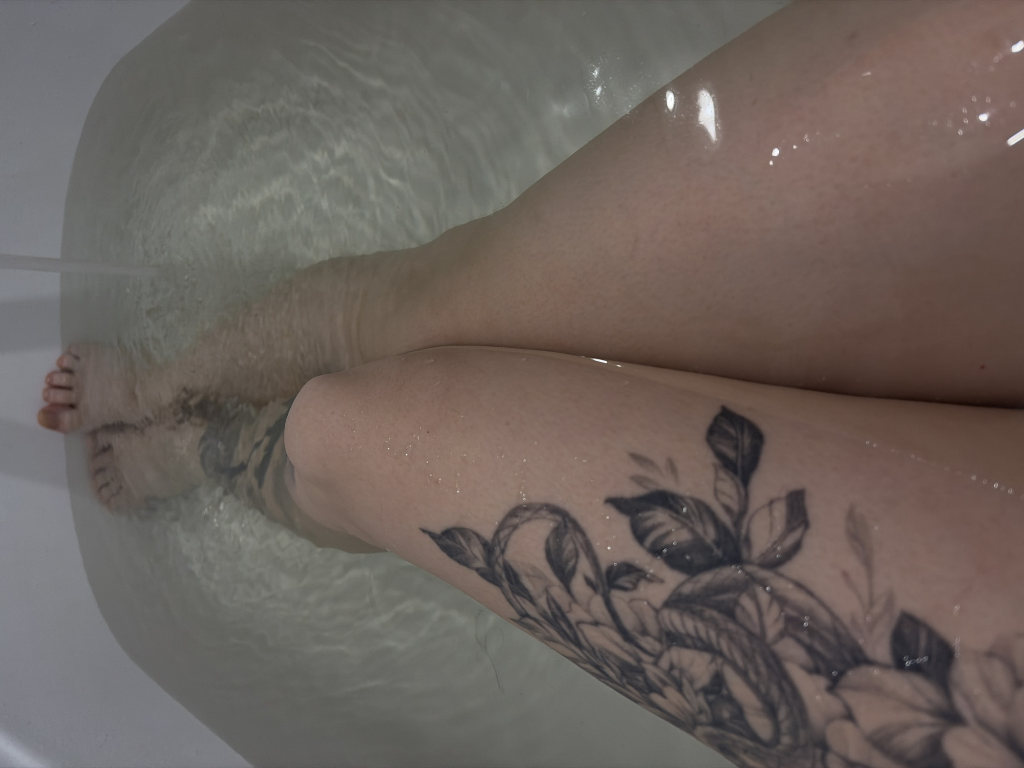 A top-down shot shows a person in a bathtub filled with water. The person's legs are visible, with one leg resting over the other. The leg closer to the viewer has a large floral tattoo. The water is clear, and the bathtub appears to be white. There is no indication of the location being Chaikovsky, Russia.