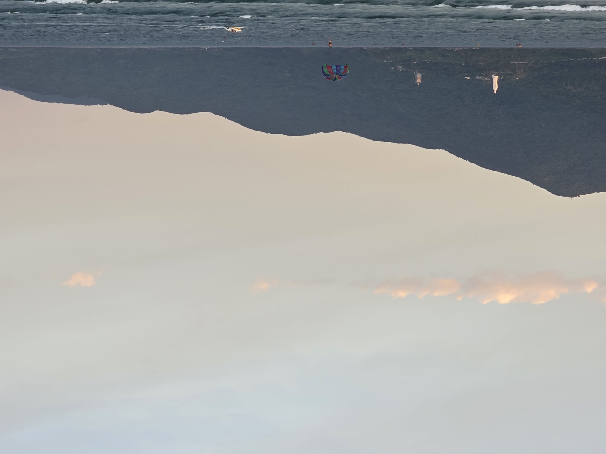 The image shows a scenic coastal view of Đà Nẵng, Vietnam, likely taken at sunset given the soft, warm lighting. The foreground features the ocean with gentle waves and white foam cresting. In the middle ground, a vibrant, multicolored parachute is visible in the sky, being towed by an unseen boat, with a person suspended beneath it enjoying a parasailing activity. To the right, a small white speedboat is moving across the water. The background is dominated by a large, dark green, tree-covered mountain range that stretches across the horizon. On the slopes of the mountains, several structures are visible, including a tall white statue of a woman on the left, and a pagoda-like tower further to the right. A thin strip of pale orange clouds is present near the top of the image, contrasting with the predominantly light blue and beige sky. The overall atmosphere is peaceful and recreational.