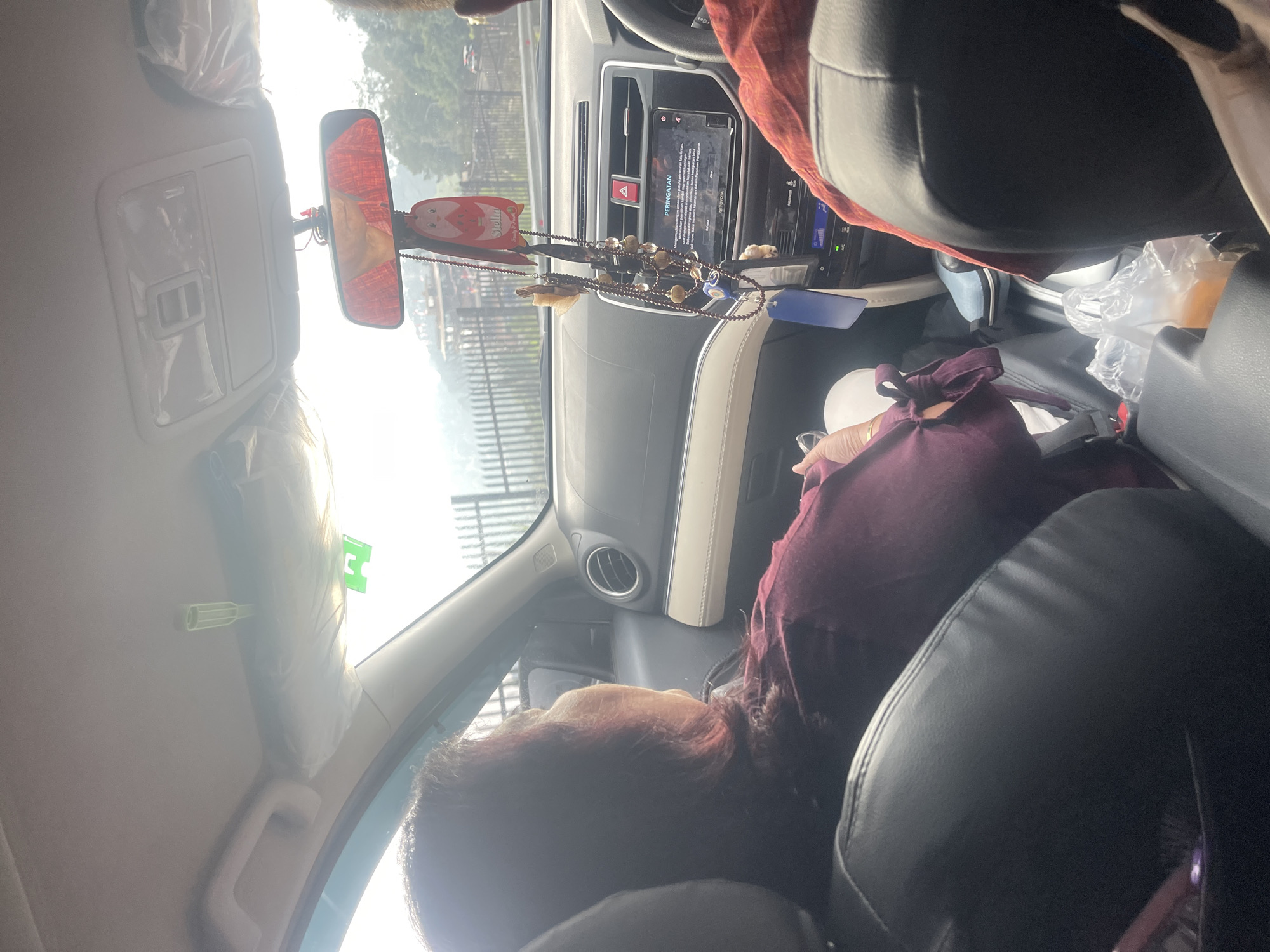The image captures the interior of a car, likely a Toyota, with two individuals visible from the chest up. The person in the foreground, facing away from the camera, appears to be a woman with dark, reddish-brown hair, wearing a maroon long-sleeved shirt with tied sleeves. She is seated in the passenger seat. In the background, another individual, possibly a driver, is partially visible, wearing a red shirt.
The car's dashboard features a central infotainment screen displaying a warning message in Indonesian, along with the Toyota logo. Various decorative items are hanging from the rearview mirror, including a red ornament with a cartoon character and what appears to be a string of beads and wooden charms. A blue tag is also hanging. Sun visors are present on the ceiling, with one covered in plastic. A vent is visible on the right side of the dashboard.
Outside the car, through the windows, a blurred view of a fence and trees can be seen, suggesting a natural or semi-rural setting. The sky is bright and appears to be daytime, with sunlight casting glare on the interior. Based on the context of the warning message on the screen and common locations for such scenes, the car is likely in Cisarua, Indonesia, a popular tourist area known for its natural beauty and winding roads. Plastic bags containing food or drinks are visible on the floor between the seats.