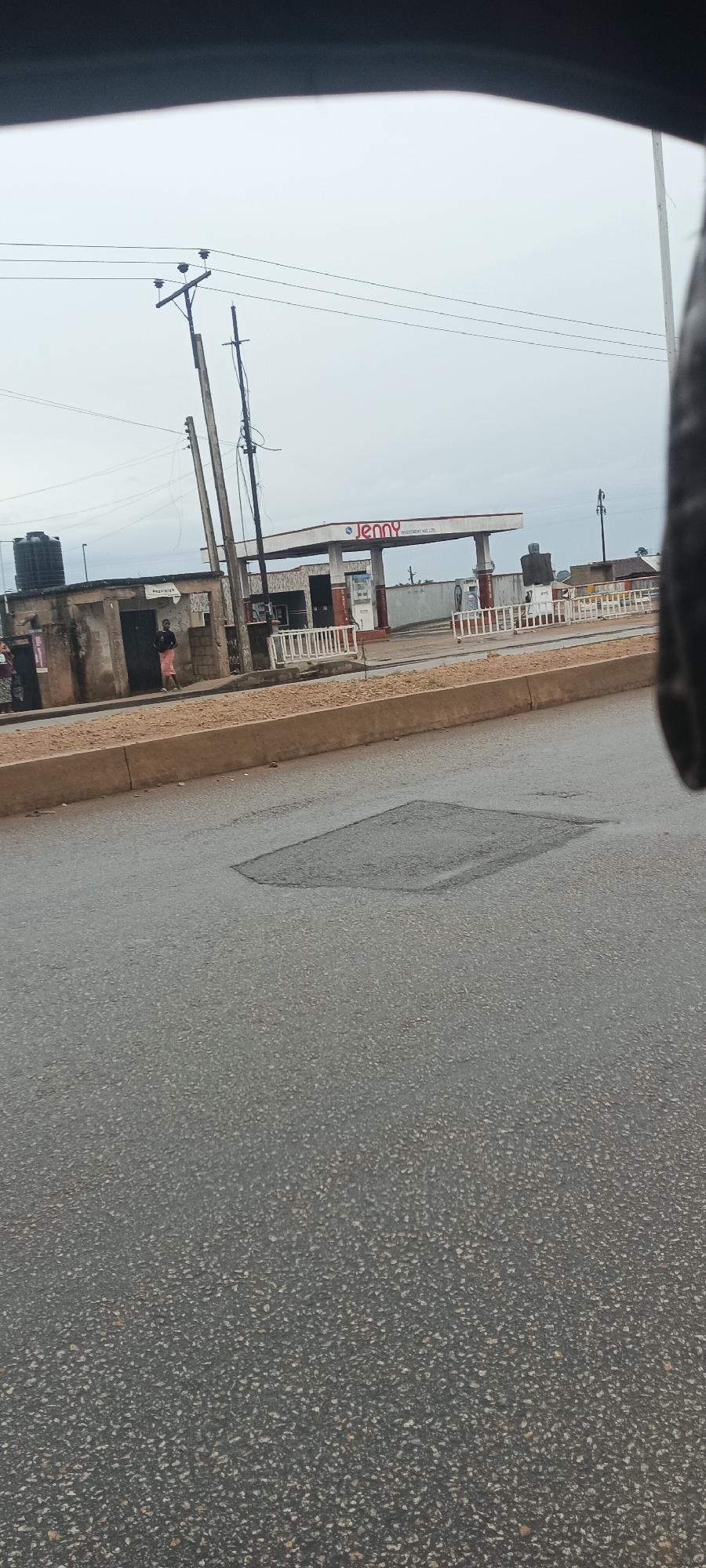 The image presents a roadside view from within a vehicle on an overcast day in Bukuru, Nigeria. The foreground shows a grey asphalt road with visible aggregate and a distinct rectangular patch, indicating road repair or a pothole. A low concrete curb separates the road from a dirt shoulder.
In the midground, a "Jenny" gas station is the most prominent feature, identifiable by its red sign that reads "Jenny Investment Nig. Ltd." The station has a canopy covering fuel pumps, and a white barrier or fence is situated in front of it. To the left of the gas station are several older, concrete-block buildings. One of these structures has a small sign near its entrance that appears to say "PRECISION". A large black water tank is visible on the roof of a building further to the left.
Several utility poles with overhead power lines stand along the roadside in front of these buildings, leading up into the grey, cloud-covered sky. A few individuals are visible near the buildings to the left of the gas station; one person, wearing light shorts and a dark top, stands near the "PRECISION" sign. The overall sky is uniformly grey and overcast, suggesting a cloudy day with muted lighting. The perspective is from a moving or stationary vehicle, with part of the vehicle's interior visible at the top of the frame.