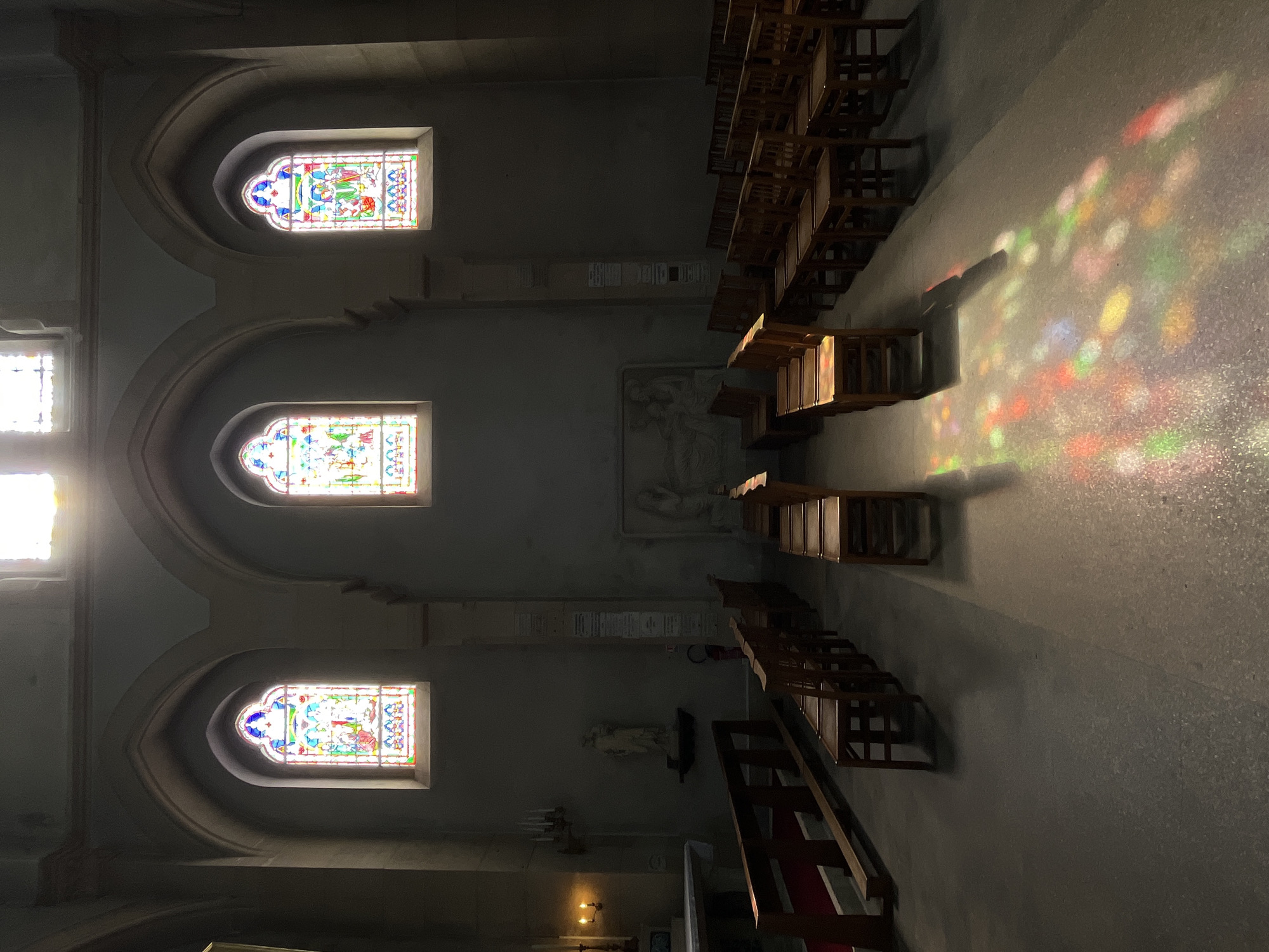 The image depicts the interior of a church, likely the Église Saint-Lubin in Rambouillet, France, due to the architectural style and the presence of stained-glass windows. The scene is dimly lit, with shafts of colorful light from three arched stained-glass windows illuminating the otherwise somber interior. Rows of simple wooden chairs are arranged on the floor, with some bathed in the colored light.
In the foreground, wooden pews with red carpeting are visible on the left. A small altar area with a white surface and a crucifix is also present. To the left of the altar, a statue of a figure in a white robe stands on a shelf. Further back, a bas-relief carving is mounted on the wall. The walls are a muted grey, and the ceiling features arched structures above the windows. The overall impression is one of quiet reverence and solemnity.