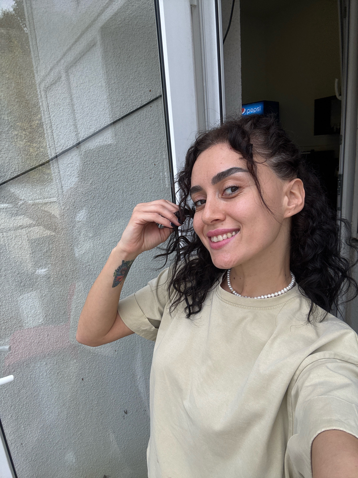 A young woman with dark, curly hair and a pearl necklace poses for a selfie in front of a textured, light-colored wall, possibly outside a building. A glass door or window reflects a tree and building. To the right, a small blue Pepsi dispenser is visible. The woman is wearing a light beige t-shirt and has a colorful tattoo on her left forearm. She is smiling at the camera and touching her hair with her right hand. The scene appears to be located in Tbilisi, Georgia, based on the visual cues and context provided.