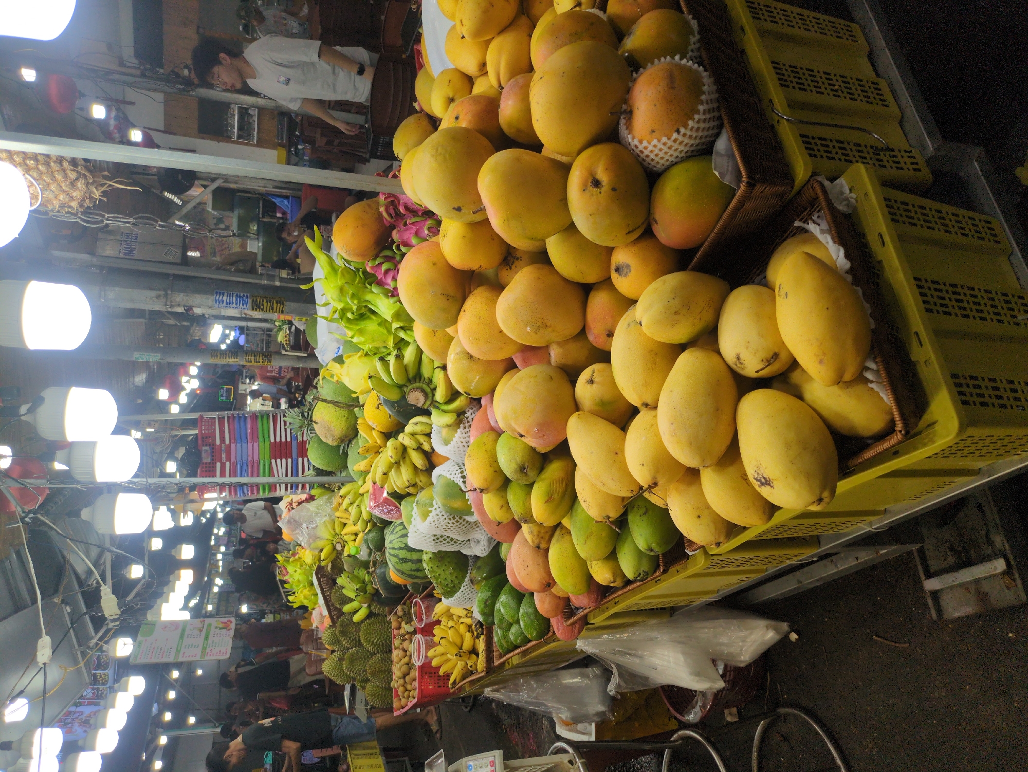 The image depicts a vibrant fruit stall in a market in Phú Quốc, Vietnam. Large piles of yellow mangoes are stacked high in woven baskets placed on yellow plastic crates. Additional fruits, including bananas, watermelons, durian, and possibly dragon fruit, are visible. Fluorescent lights illuminate the scene. People, including a vendor in a white shirt, are present in the background, suggesting the market is active. Other market stalls and signage are partially visible. The ground appears to be dark and slightly wet.