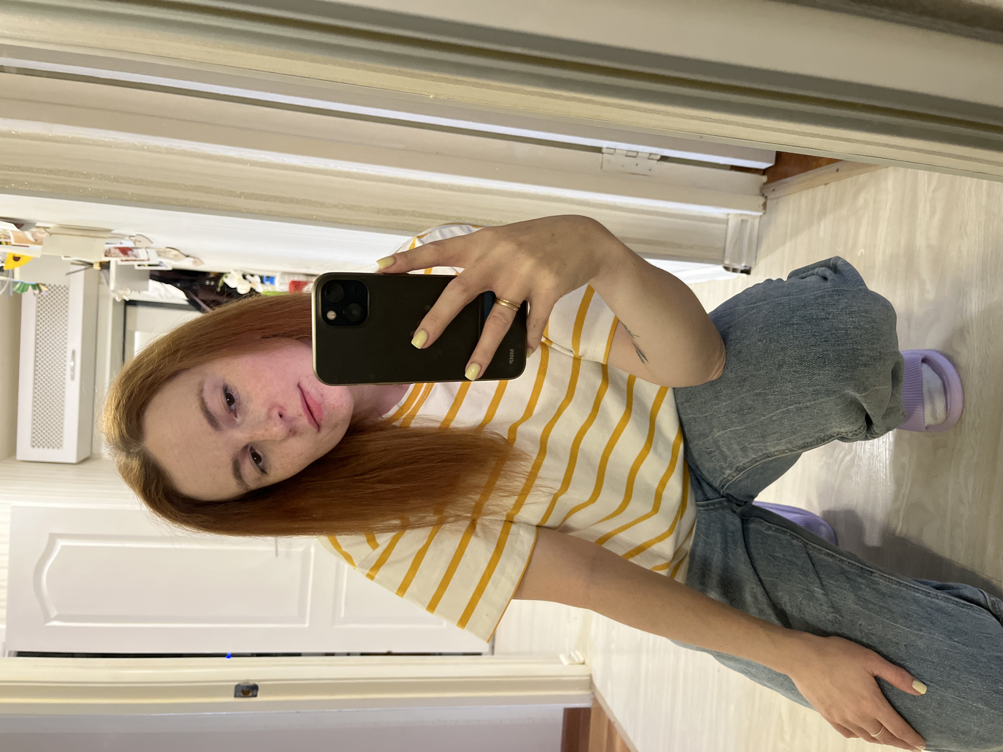 A young, fair-skinned person with reddish hair and visible freckles poses for a selfie in a mirror. They are wearing a white and yellow striped t-shirt, blue jeans, and light purple socks. Their fingernails are painted yellow. They are holding a black iPhone in their right hand, and their left hand rests on their left leg. A gold ring is visible on their right ring finger.
The reflection shows a room with light-colored walls and flooring. A white door and various objects are partially visible in the background.
Given the request, it is not possible to independently verify the location as Chaikovsky, Russia, without additional information or identifiers present within the image.