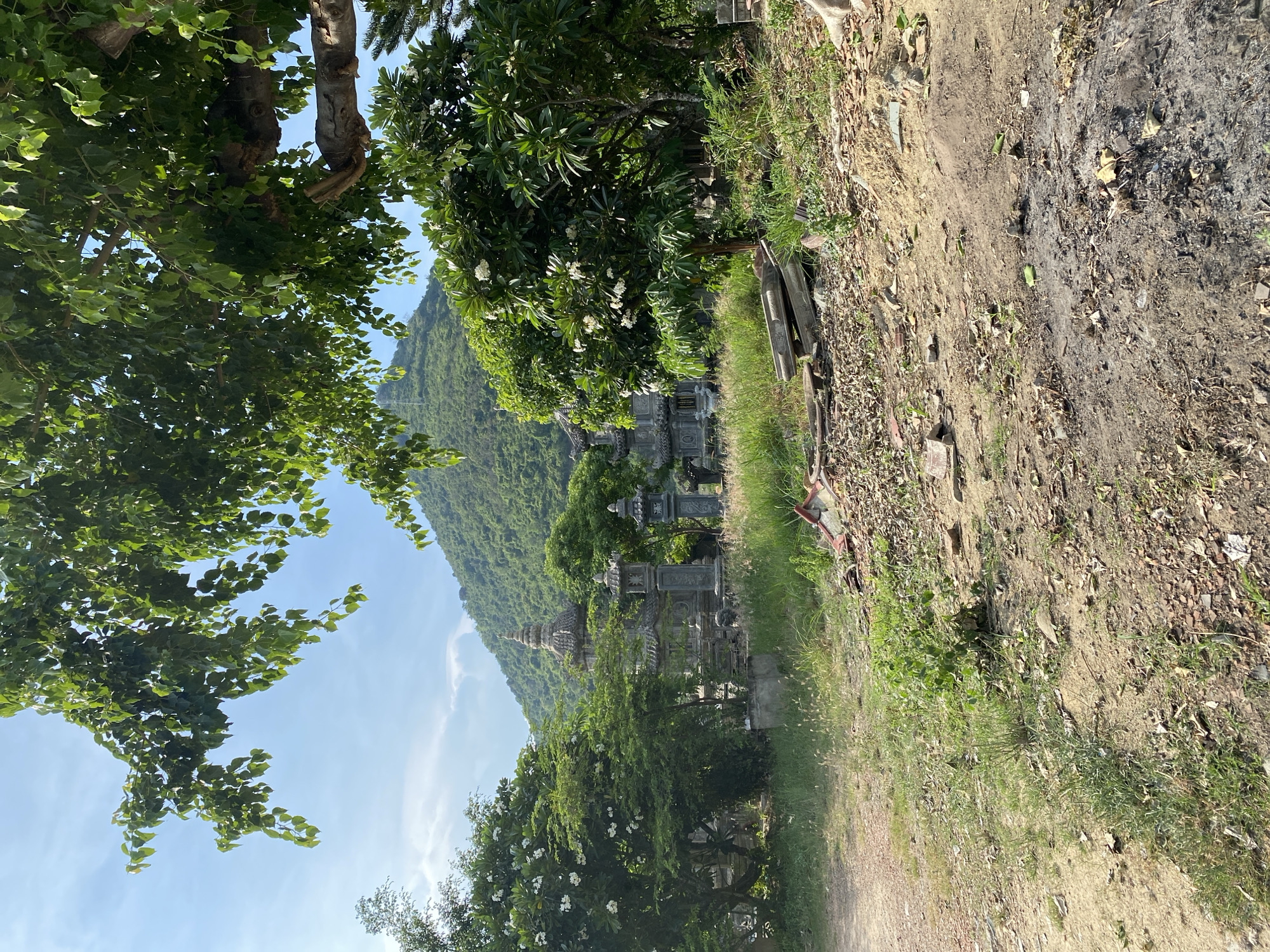 The image depicts an outdoor scene with a verdant, tree-covered mountain in the background under a partly cloudy sky. In the mid-ground, there are several ornate stone structures, resembling small temples or monuments, surrounded by lush greenery and flowering trees. The foreground is a dusty, uneven ground with scattered debris, including broken pieces of red tiles and dry leaves. A dirt path is visible to the left. The overall impression is of a temple complex or a historical site situated in a natural, hilly landscape. The specific location mentioned, Tay Nha Trang Ward, Vietnam, suggests a tropical or subtropical environment.