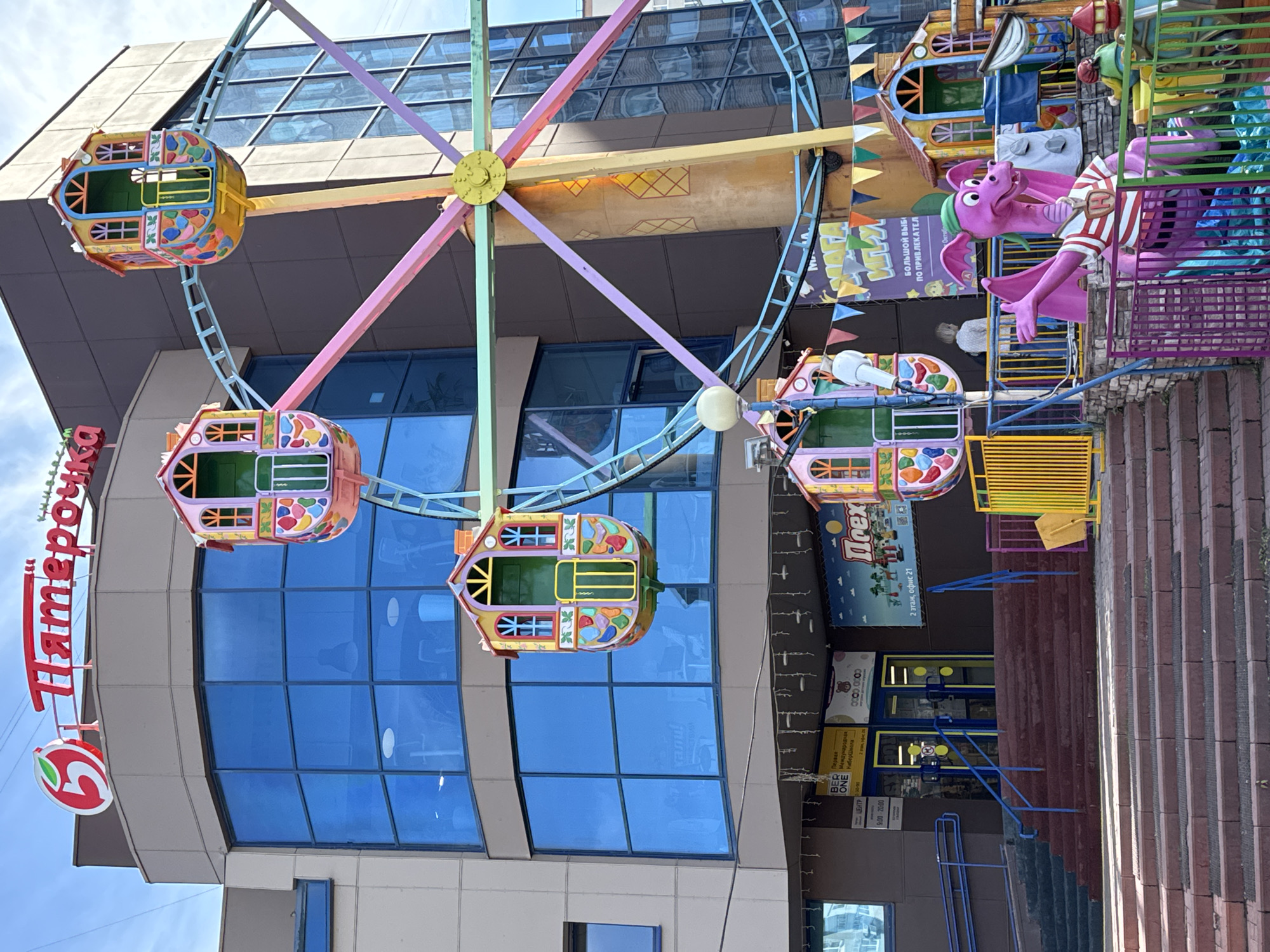 The image captures a colorful amusement park scene in an urban setting, specifically in front of a building with a prominent "Пятерочка" (Pyaterochka) sign, a Russian chain of grocery stores. This suggests the location is likely in Russia.
The main subject is a miniature Ferris wheel, featuring brightly colored carriages resembling small houses. The Ferris wheel is decorated with rainbow-colored spokes. The foreground also shows a ride with a purple dragon figure.
Behind the Ferris wheel, the modern building is characterized by a facade with large blue-tinted windows. Additionally, a poster or sign for "Прех" is visible, and a door is accessible by a staircase. The stairs appear to be made of red brick or paving stones.
The sky is visible in the upper left corner, indicating it is a daytime shot. The image is taken from a low angle, looking upwards towards the Ferris wheel and the building.