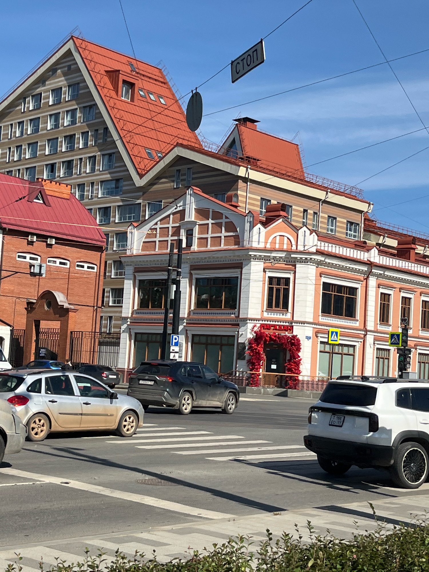 The image captures a street scene in Perm, Russia, likely at an intersection, with several buildings and vehicles visible. Dominating the background is a multi-story building with a distinctive steep, orange-tiled roof, characteristic of some modern architectural styles in the city. Adjacent to it is another building with a red tiled roof and brickwork, followed by a larger, ornate building with a light-colored facade featuring white trim and dark windows. A traffic light and street signs, including a "STOP" sign hanging overhead, are present. In the foreground, several cars are seen on the road, some stopped at a pedestrian crossing. A silver hatchback, a dark SUV, and a white SUV are the most prominent vehicles. The environment suggests a daytime scene under a clear blue sky.