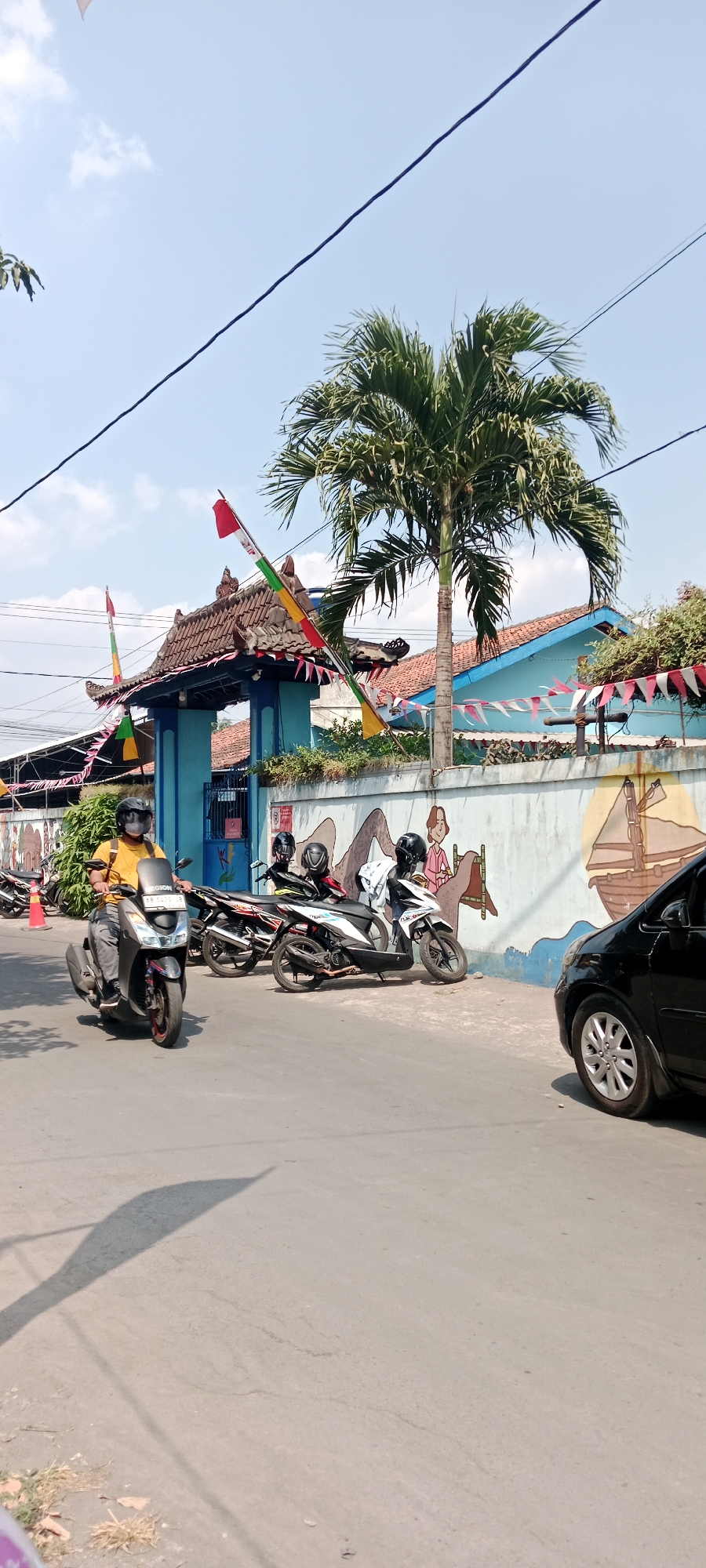 In Purwomartani, Indonesia, a street scene unfolds under a bright, clear sky. A person rides a dark-colored scooter, wearing a yellow shirt and a black helmet, moving from left to right in the foreground. Behind them, parked on the side of the road, are several other scooters. A black car is visible on the right side of the image. The background features a blue building with a traditional tiled roof, adorned with red, white, and green flags and pennants, suggesting a celebration or holiday. A palm tree stands tall near the building, and a mural depicting figures and a sailing ship decorates the light-colored wall. Electrical wires crisscross the sky above the scene.