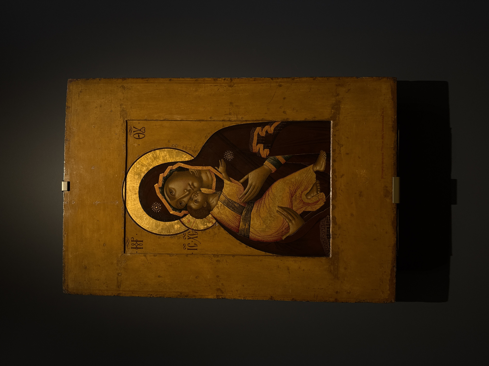 The image shows an Orthodox icon of the Virgin Mary holding the infant Jesus. The icon is painted on a rectangular wooden panel with a gold-colored frame. The faces of Mary and Jesus are depicted with large, almond-shaped eyes and long, narrow noses. Mary is wearing a dark brown head covering and robe. Jesus is wearing a yellow garment. A gold halo surrounds the heads of both figures. There are Greek letters on the left and the right sides of the halo. The icon is mounted on a dark wall, likely within a museum or gallery setting. The location of the icon is Moscow, Russia.