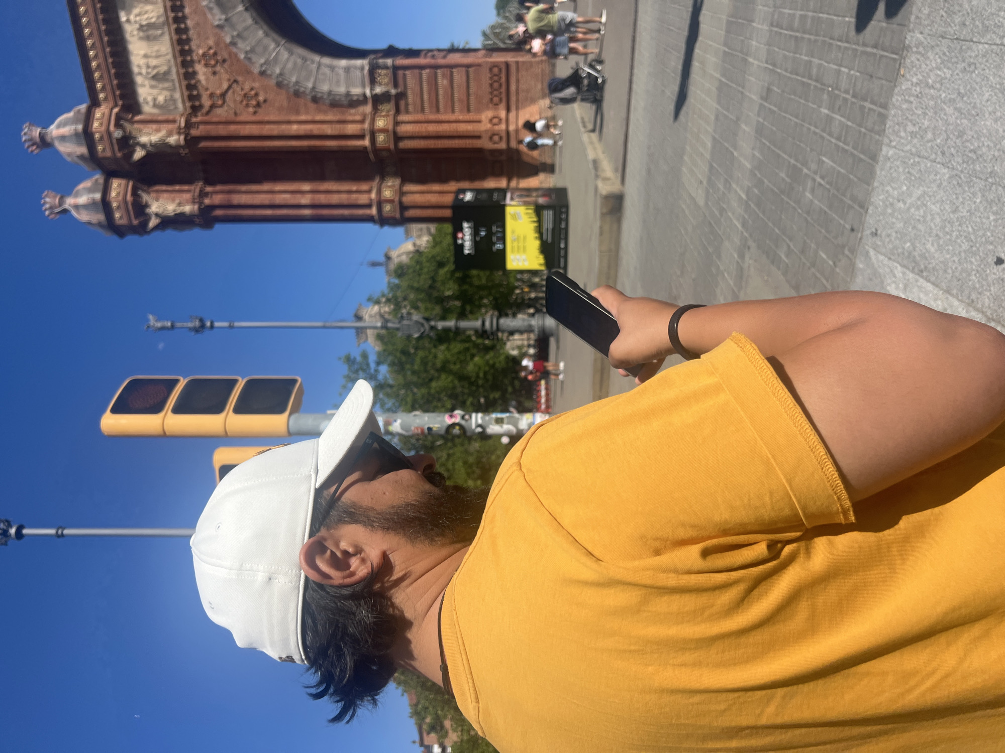 The image shows a person wearing a yellow t-shirt and a white baseball cap, holding a black smartphone. In the background, the Arc de Triomf in Barcelona, Spain is visible, along with a traffic light, trees, and other people on the sidewalk.