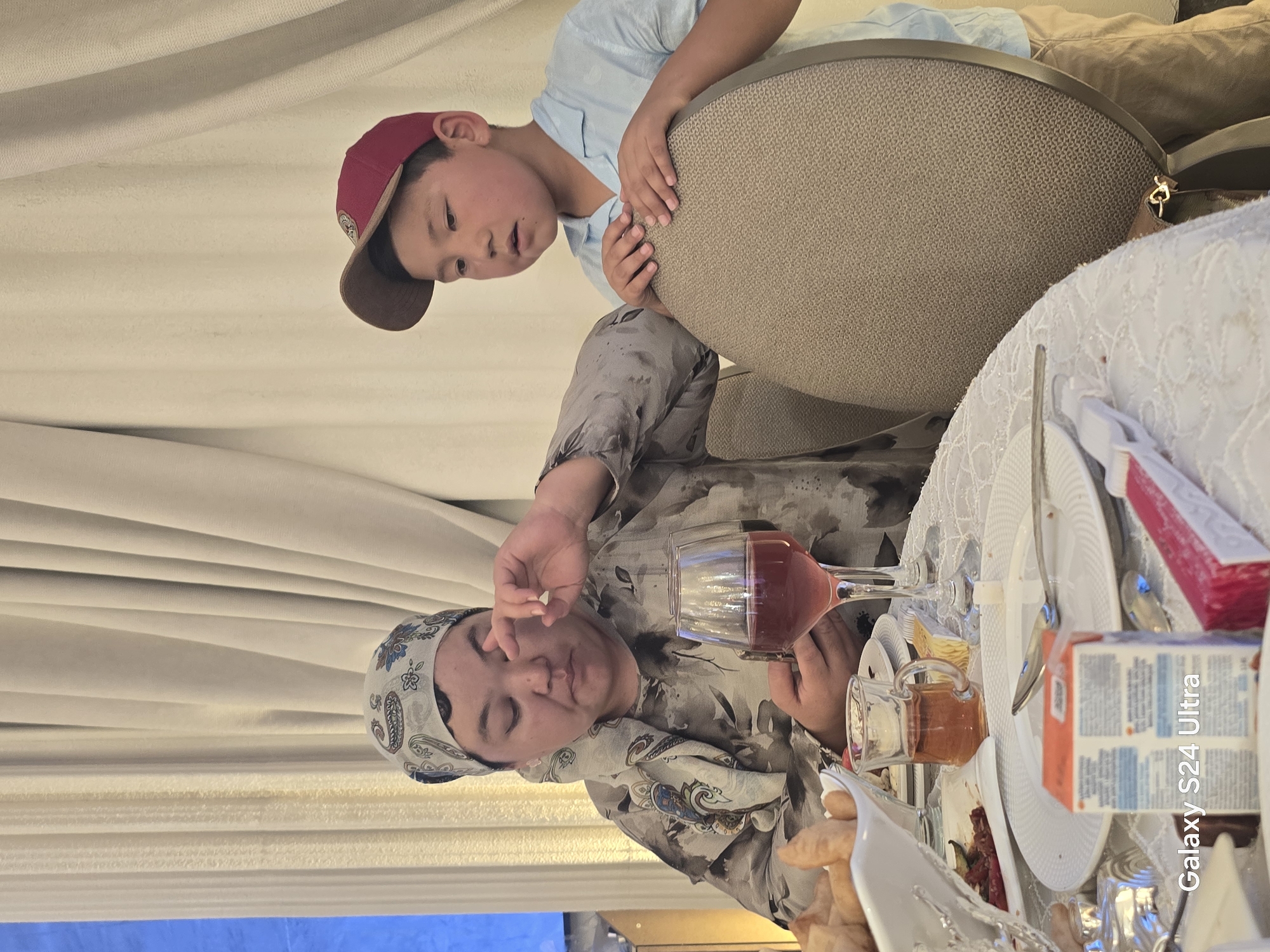 In Karakol City, Kyrgyzstan, a woman wearing a patterned headscarf and a printed shirt is seated at a table. She appears to be interacting with her eye, possibly with a finger. To her right, a young boy in a red baseball cap and a light blue shirt stands leaning on a chair. The table is set with plates, cutlery, a glass of red liquid, and a small glass of amber-colored liquid. There is also a white bowl containing food, and a small juice box is visible in the foreground. The background consists of draped curtains.