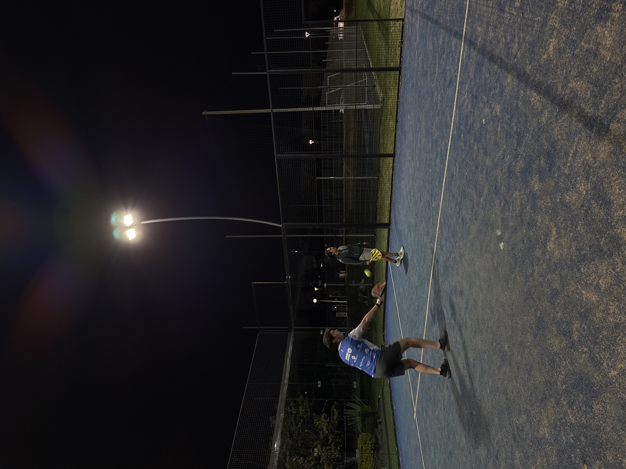 The image depicts a nighttime scene on a blue paddle tennis court in Heroica Puebla de Zaragoza, Mexico. A man in a blue shirt and dark shorts is in the process of swinging a paddle tennis racket at a yellow tennis ball. Another individual stands near the far end of the court holding a racket. The scene is illuminated by overhead artificial lighting on a pole, with darkness surrounding the area beyond the court. A mesh fence encloses the court.