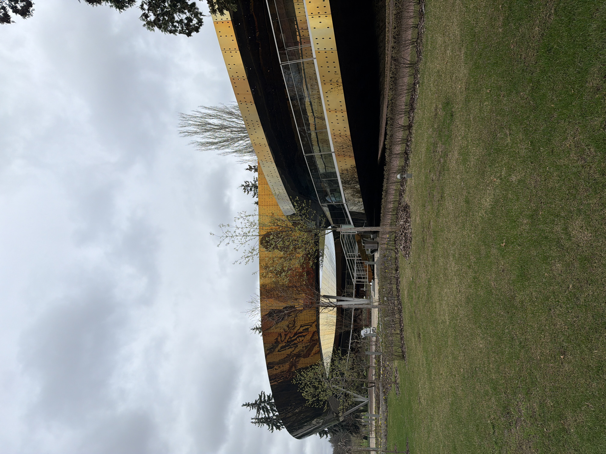 The image depicts an exterior view of the Garage Museum of Contemporary Art, located in Moscow, Russia. The structure is characterized by a curvilinear facade clad in reflective metal panels. The building's design integrates glass elements, allowing for interior illumination. The surrounding landscape features a green grassy area and trees with budding foliage, suggesting a springtime setting. The sky is overcast, casting a diffuse light over the scene. No people are visible in the frame.