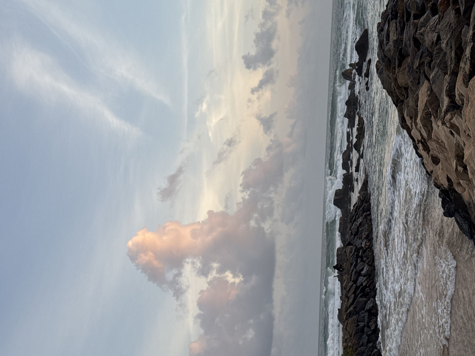 This media file captures a scenic view of a beach with rocky outcrops and a turbulent sea, under a sky filled with dramatic clouds. The scene is oriented vertically, giving a sense of looking up at the sky or down a steep incline.
The primary subjects are the natural elements: a rough, dark-colored rocky coastline occupies the right side of the frame, with waves crashing against it. The ocean stretches out to the left, showing a dynamic interplay of white foam and greenish-blue water. Above the sea and rocks, the sky is a spectacle of clouds. There are wispy, pale clouds near the top, and more substantial, darker clouds interspersed with patches of light, some tinged with pink and orange, suggesting the soft light of either sunrise or sunset.
A single human figure is visible, perched on the rocks near the water's edge, appearing small against the vastness of the natural landscape. This suggests a moment of quiet observation or contemplation within the powerful environment.
The time of day is likely during the "golden hour" due to the warm hues in the clouds, implying either early morning or late evening. The weather appears to be dynamic, with visible waves indicating possibly windy conditions or a strong tide. The location is identified as Midigama, Sri Lanka, which is known for its beaches and surf spots, aligning with the imagery of the ocean and rocky shore.
There is no visible text in the media file. The overall mood conveyed is one of awe at nature's power and beauty, with a sense of solitude and introspection.