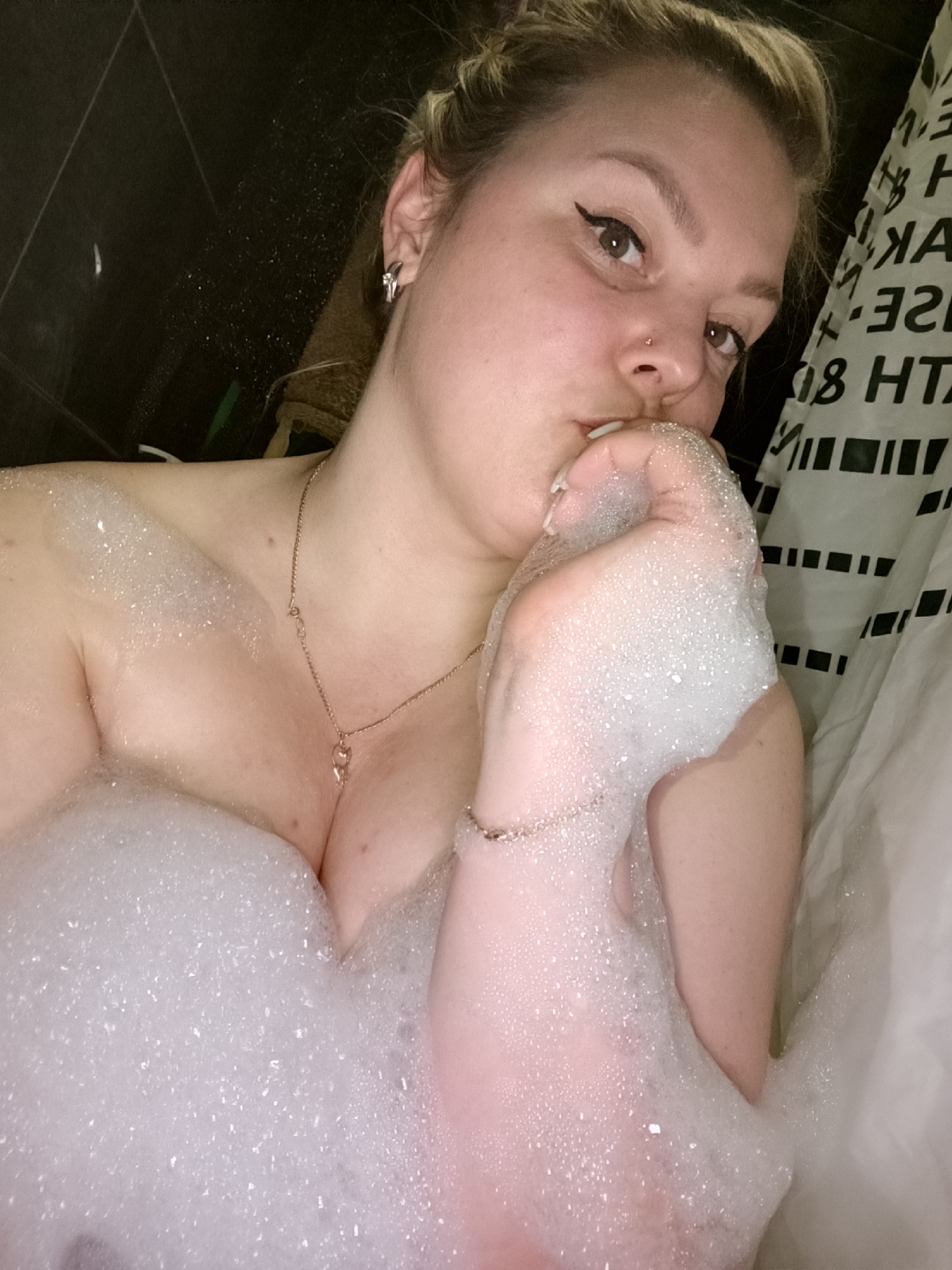 A young woman is captured in a close-up, high-angle shot, enjoying a bubble bath. Her upper body is partially submerged in a mass of white, foamy bubbles. She has light-colored hair pulled back and is looking directly at the camera with a neutral expression. Her visible left eye is accented with winged black eyeliner. She wears a small gold stud nose piercing, a silver-toned huggie or cuff earring with a small stone on her right ear, a thin gold chain necklace with a small pendant (resembling a cat or heart), and a thin bracelet on her right wrist.
The woman is seen holding a large clump of bubbles to her mouth, appearing to blow or kiss them playfully. The setting is a bathroom, with a dark, possibly tiled wall visible in the background on the left. On the right, a white shower curtain with a distinctive pattern of black horizontal rectangular bars and stylized, reversed text is visible. Portions of the text read "E H", "XA", "ИАЗ", "3E", and "08 HT", suggesting either Cyrillic letters or abstract decorative text. The lighting is artificial, indicative of an indoor setting, likely in the evening or at night. The activity is a relaxing bubble bath. While the provided context specifies Марковский, Russia, there are no explicit visual cues within the image itself to confirm this specific location.