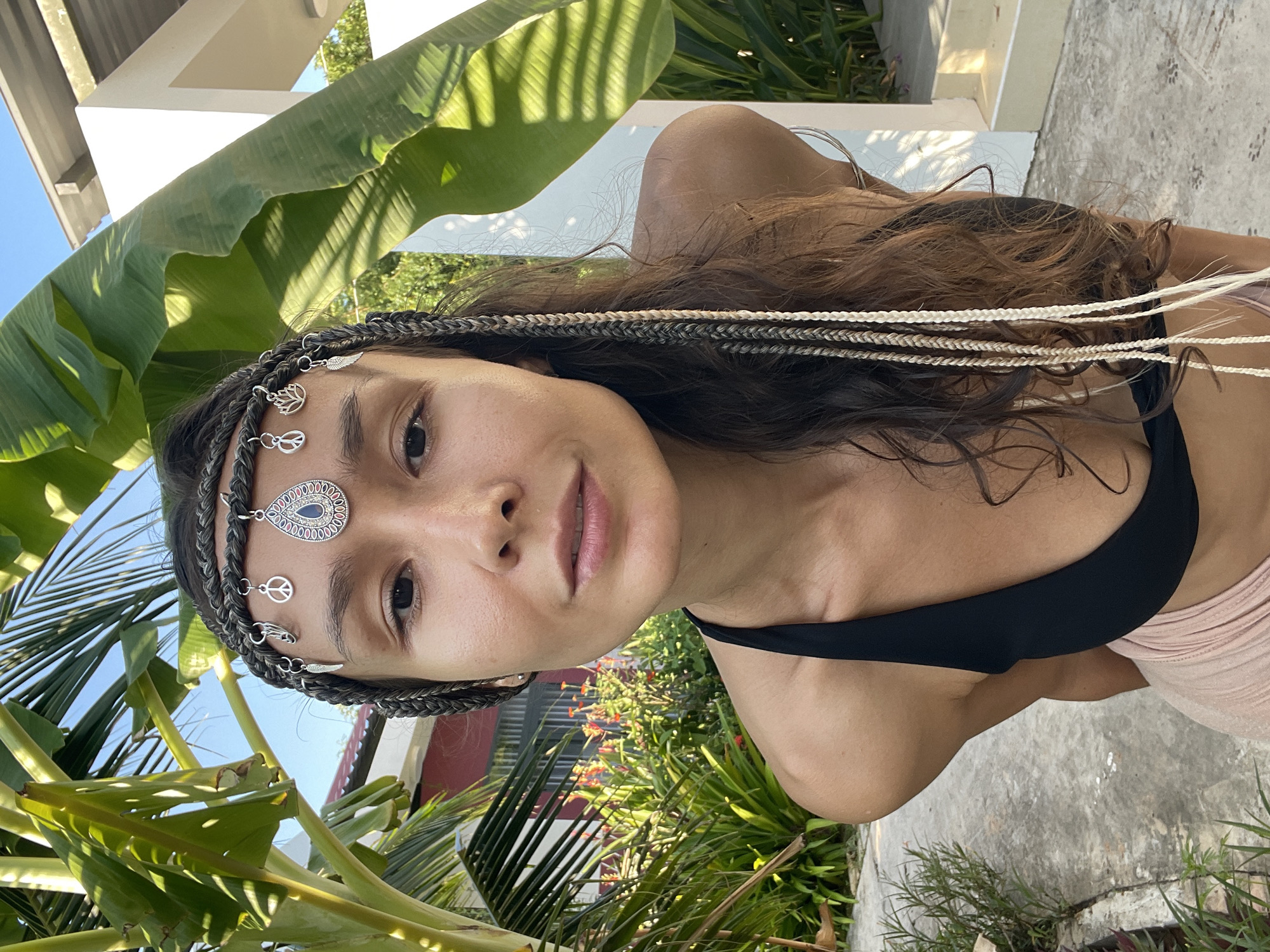 A young woman with dark hair and a bohemian-style headpiece adorns her forehead. The headpiece consists of braided dark material, adorned with small silver charms in the shape of a peace sign, a lotus flower, and a feather, as well as a larger teardrop-shaped pendant with blue and silver detailing. She is wearing a black bikini top and a light pink skirt. The woman is positioned outdoors, leaning forward and looking directly at the camera. Behind her, lush green foliage, including large banana leaves and palm fronds, creates a vibrant backdrop. In the distance, a building with a red facade is visible, along with a white structure. The ground beneath her appears to be paved with concrete. The lighting suggests a sunny day. The location is identified as Phu Thuy Ward, Vietnam.