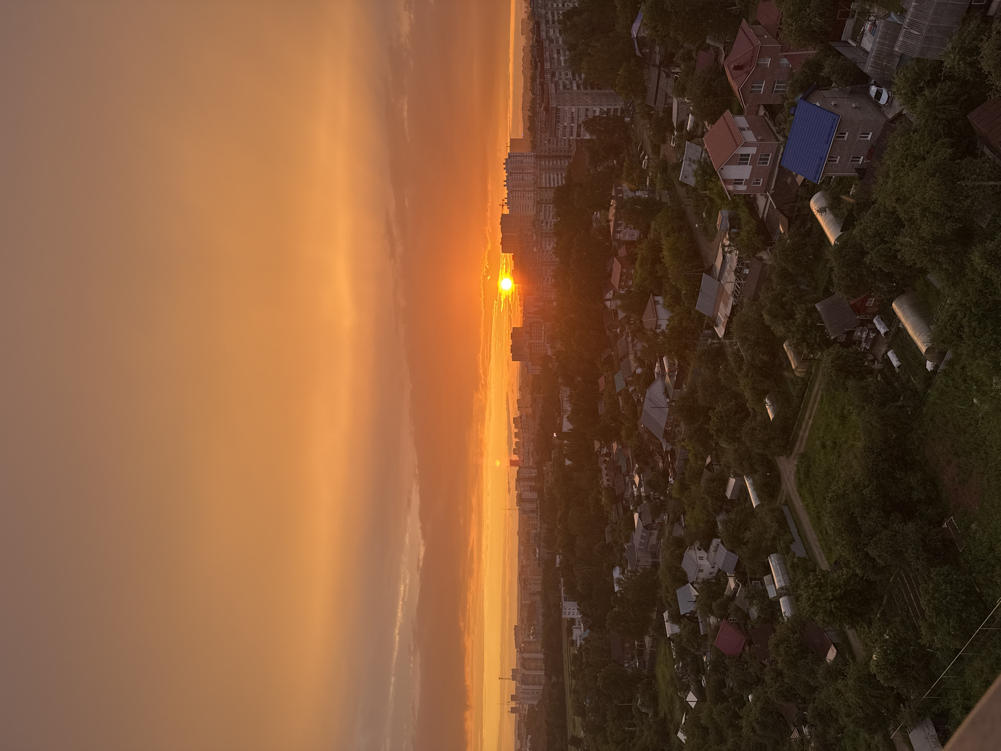 The image depicts an aerial view of a city at sunset. The sun, a bright orange orb, is low on the horizon, casting a warm glow over the scene. The sky is filled with hues of orange and yellow, with some faint clouds. To the left of the frame, the sky dominates, with the sun's rays creating a soft, diffused light.
The right side of the image shows a cityscape transitioning into a more suburban or rural area. In the foreground, there are numerous houses with red and blue roofs, nestled among lush green trees. Some of these houses appear to be small residential buildings, while others are larger, multi-story structures. There are also cylindrical tanks, possibly for water or industrial use, scattered throughout the area. Roads and pathways wind through the dense foliage and buildings.
In the distance, a line of taller apartment buildings marks the edge of the city. The overall impression is one of a peaceful evening in a Russian city, likely Izhevsk, given the context. The warm sunset colors evoke a sense of calm and tranquility. The activity taking place is simply the passage of time, as the day ends and the city prepares for night. No people are explicitly visible, but the presence of houses and buildings suggests human habitation. There is no visible text in the image. The weather appears to be clear, with the sunset being the prominent atmospheric feature.