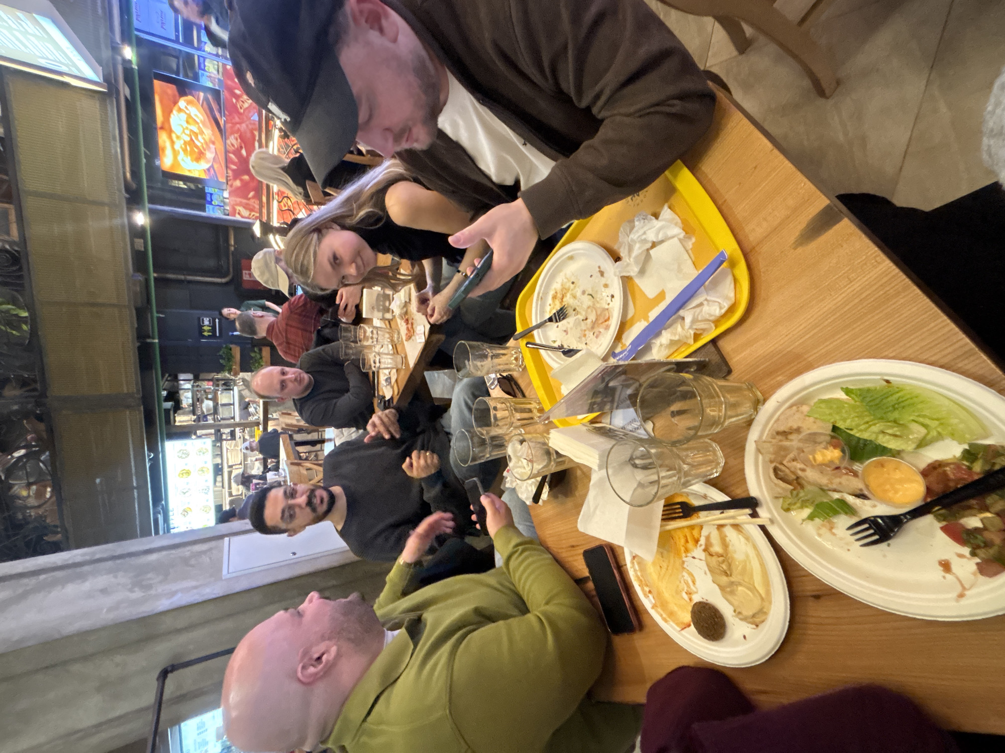 A group of people is seated around a wooden table in a restaurant. Empty plates of food are visible on the table, along with multiple glasses and silverware. Some individuals are using their mobile phones, while others are engaged in conversation. The restaurant has a modern industrial aesthetic, with exposed concrete and metal elements. A menu board and a television screen displaying images of food are visible in the background. Based on available information, it is not possible to independently verify if the location is indeed Moscow, Russia.