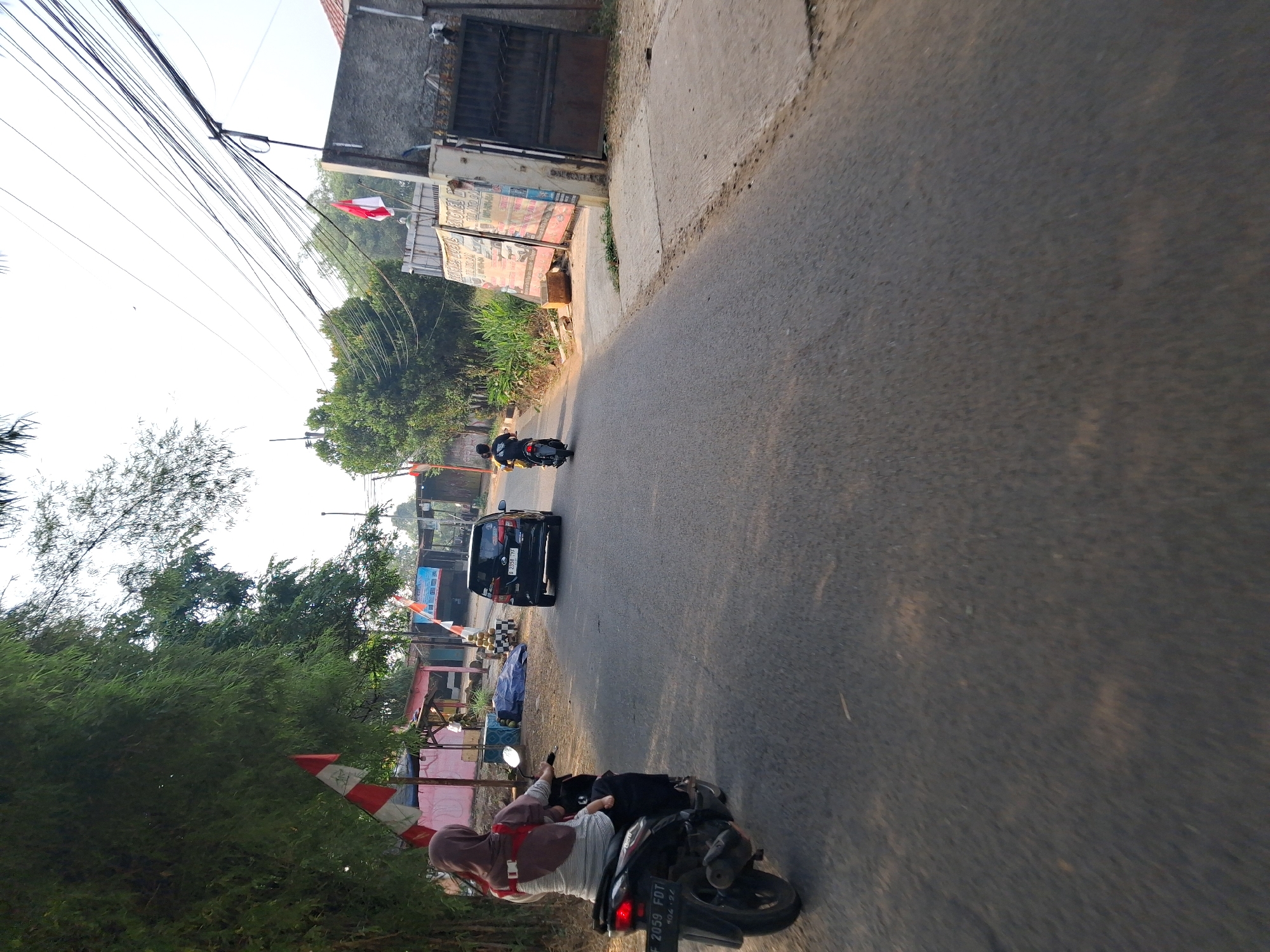 The image depicts a street scene in Bojong Gede, Indonesia, under a bright, overcast sky. A paved road, worn and with scattered debris, runs through the frame. On the left side of the road, a woman wearing a hijab and a striped shirt rides a motorcycle with a child on her lap, also wearing a helmet. The motorcycle's license plate is visible as "F 2059 FDT". Further down the road, a black car with license plate "B 2358 CYF" is driving away from the camera. Another motorcycle with a rider is visible in the distance, also moving away. The roadside features lush green foliage, including bamboo trees on the left, and some buildings with red and white Indonesian flags. On the right, a concrete wall and a metal gate enclose a property, with signs posted on the wall. Power lines are prominently strung across the upper portion of the image. The overall impression is that of a typical day in a suburban area of Bojong Gede.