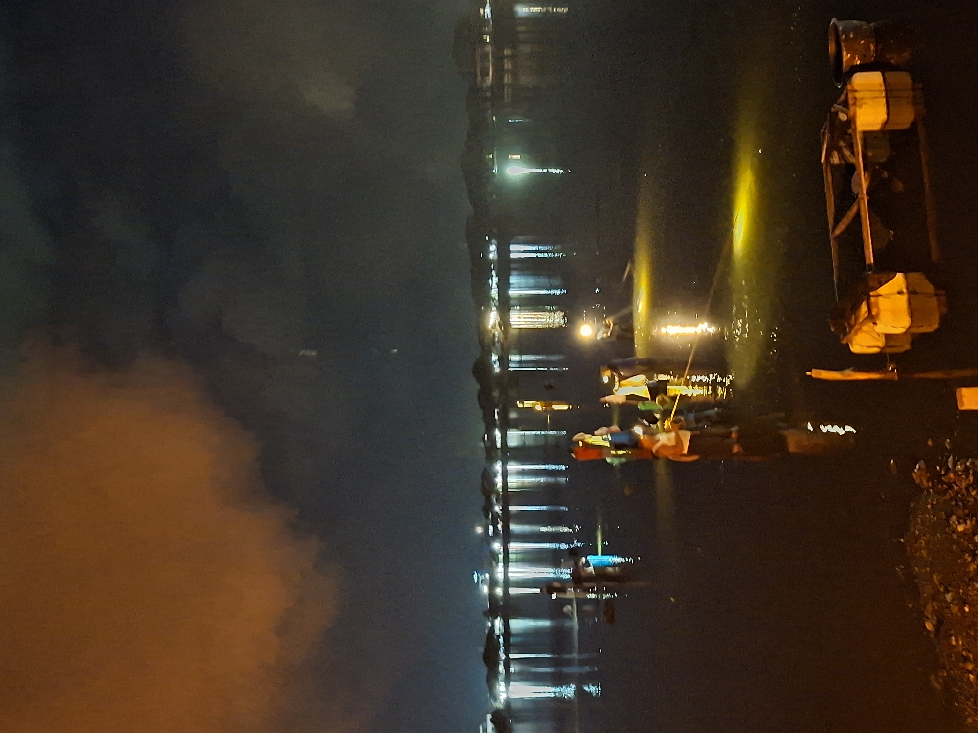 The image shows a dark, nighttime scene along the Krueng Cunda river in Lhokseumawe, Indonesia. The sky is dark and cloudy. Lights from buildings and streetlights are visible along the riverbank, reflected in the water. There are several paddle boats in the water. Some people can be seen in the boats. The water is dark with illuminated streaks from the lights. On the right side, there is a raised metal structure with orange and yellow containers. On the right edge of the image, dark foliage is visible.