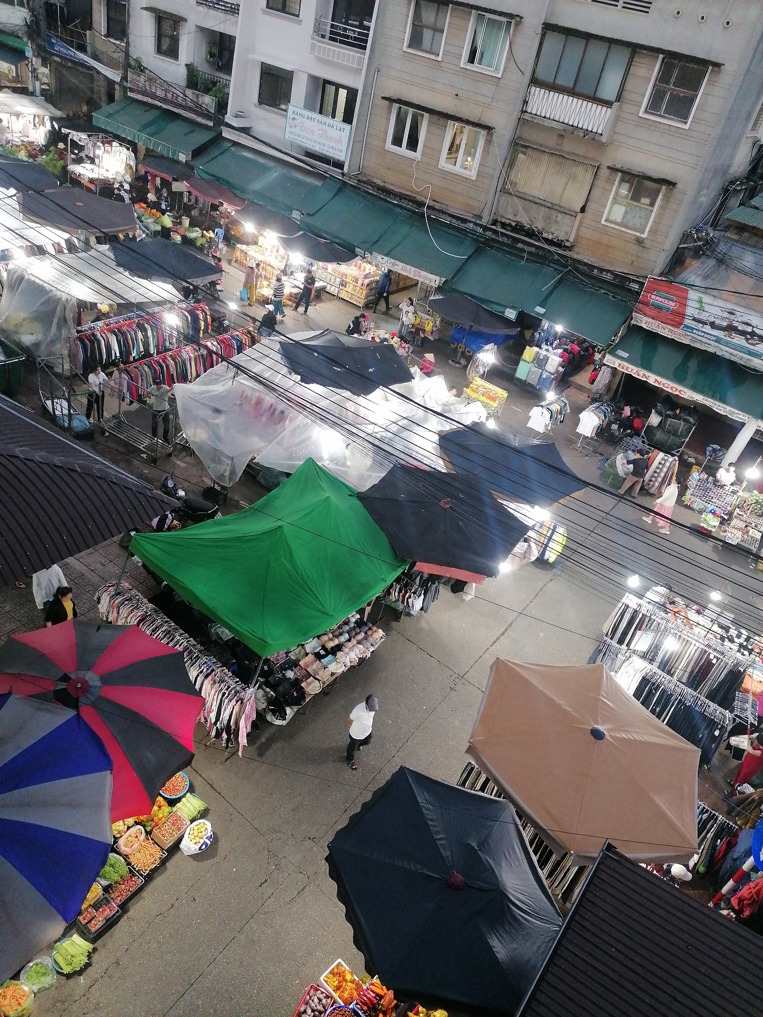 The image is an overhead shot of a bustling street market scene, likely taken from a high vantage point such as a building. The market appears to be in the city of Da Lat, Vietnam, as evidenced by the sign "HÀNG ĐẶC SẢN ĐÀ LẠT" (Da Lat Specialty Products).
The market is filled with various stalls covered by umbrellas and tarpaulins. Several stalls are selling clothes displayed on racks, with a mix of colorful umbrellas overhead, including blue/gray and red/gray patterned ones, a black one, and a tan one. There is also a stall covered by a large green tarpaulin. Some stalls have fruits and vegetables on display.
People are visible moving through the market, browsing the goods on offer. Their activities include walking, shopping, and attending to the stalls. The lighting suggests that it is either dusk or night, as the stalls are brightly lit. Buildings line the periphery of the market area, and signs are visible on their facades. Power lines are strung across the market area.