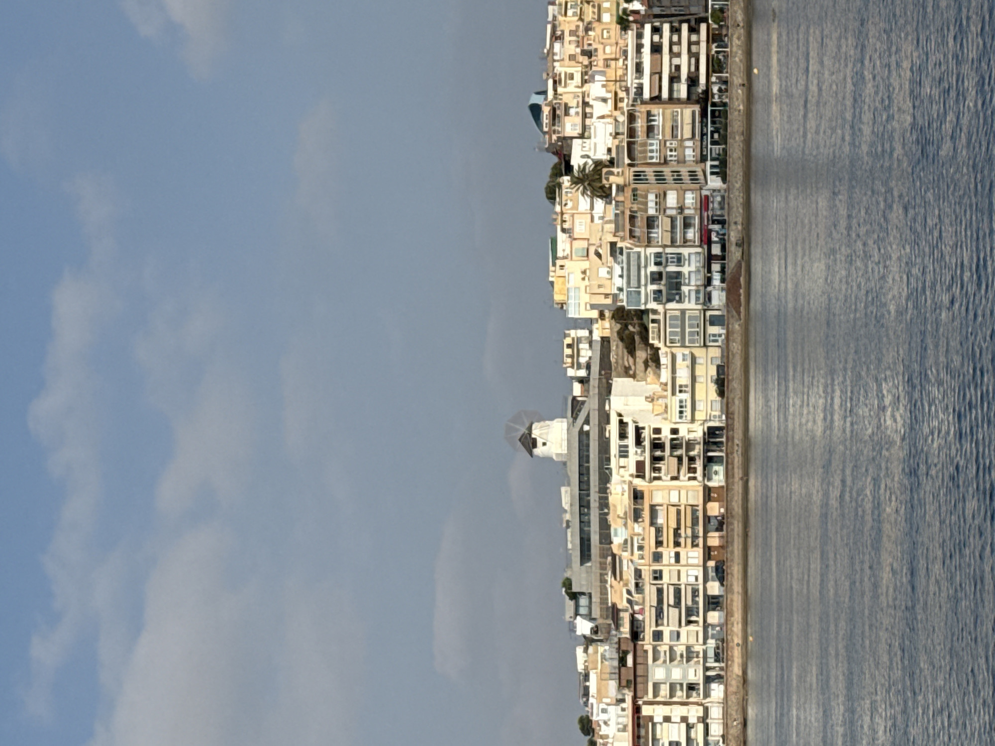 The image displays a panoramic view of the Águilas waterfront in Spain. The scene is dominated by a series of multi-story buildings lining the shore, with a body of water in the foreground. The architecture varies, featuring a mix of residential apartments and businesses on the ground floor. A prominent white windmill, a historical landmark, is visible on a hilltop behind the buildings, adding a distinctive feature to the skyline. The sky above is blue with scattered white clouds, suggesting fair weather. The water exhibits ripples, indicating a gentle movement. The buildings are painted in various light colors, including white, yellow, and beige, common in Mediterranean coastal towns.