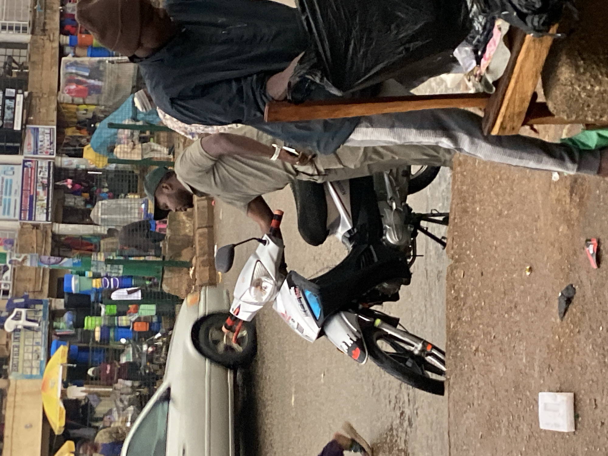 The image depicts a street scene in Bukuru, Nigeria, likely a market area, with a focus on a man on a motorcycle and other individuals. The man on the motorcycle is wearing a green cap and a light brown short-sleeved shirt, with khaki pants. He is looking to his left. Beside him, to his right, another man is partially visible, wearing a dark beanie and a dark long-sleeved shirt, holding a large black plastic bag. A silver motorcycle with black accents is prominently featured. In the background, a silver car is parked. The environment shows a busy marketplace with stalls, umbrellas, and numerous containers, possibly for sale. The ground appears wet, suggesting recent rain. The overall atmosphere is of an active urban commercial setting.