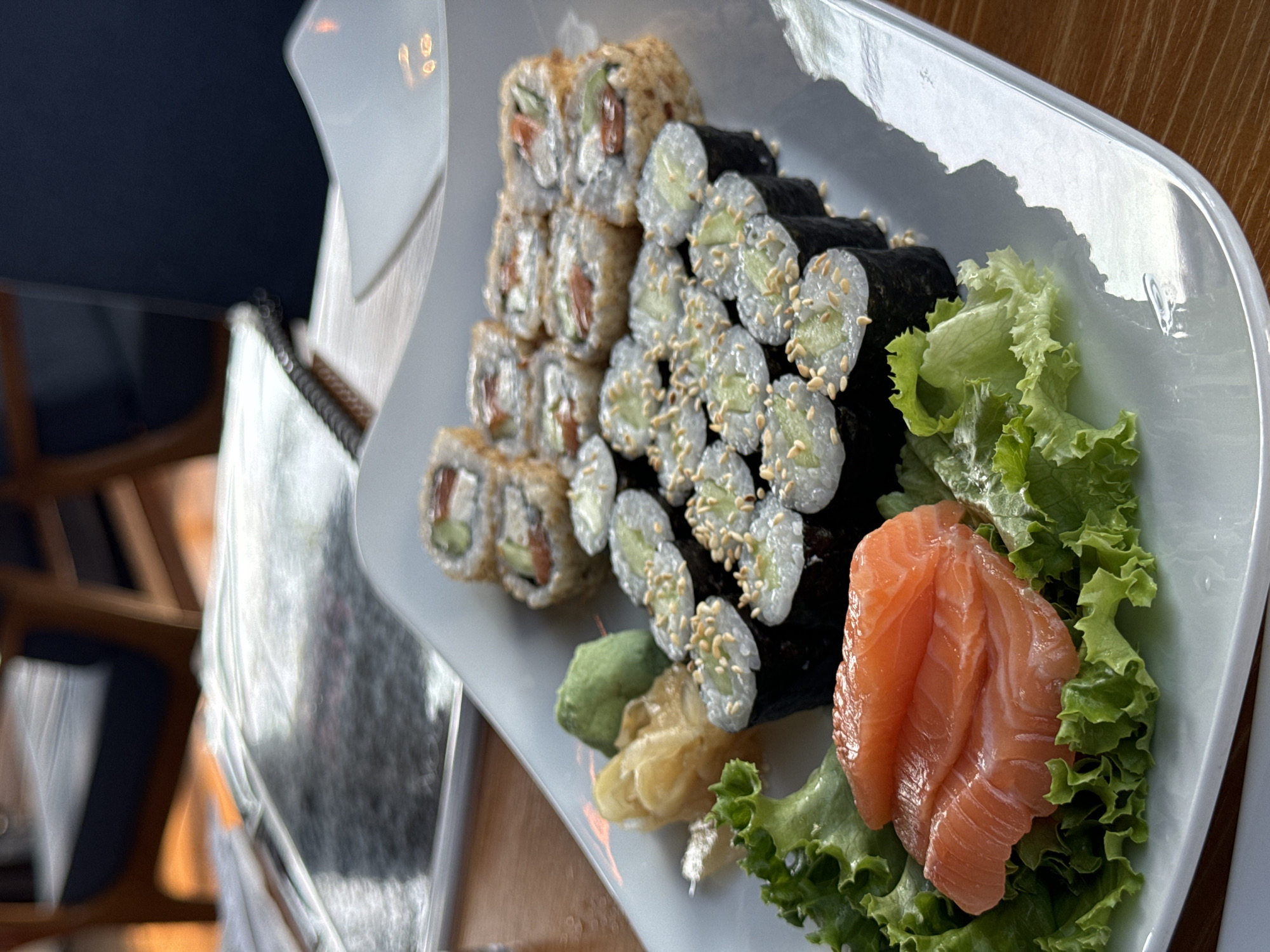 A close-up, eye-level, outdoor shot captures a white rectangular plate of sushi on a wooden table. The plate contains several pieces of sushi, including salmon sashimi, cucumber rolls topped with sesame seeds, and fried sushi rolls filled with cucumber and possibly salmon. A small pile of pickled ginger and wasabi are also present on the plate. The sushi is arranged on a bed of lettuce. In the background, a blurred view shows what appears to be a restaurant setting with chairs and a table. The lighting suggests it is daytime, with natural light illuminating the scene. No people or specific street elements are visible, preventing the precise identification of the location within Phu Thuy Ward, Vietnam.