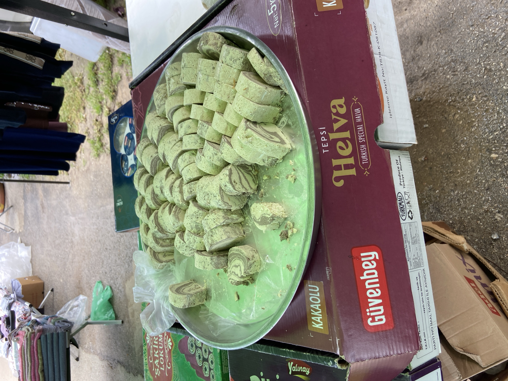 The image shows a metal tray filled with slices of halva, a Turkish sweet, placed on top of several cardboard boxes. The halva is light green in color with a swirled pattern. One of the boxes is labeled "Helva TURKISH SPECIAL HALVA" in large letters, along with the brand name "Güvenbey". Another box below the halva tray has the words "ŞAKİR LOKUM" printed on it. The setting appears to be an outdoor market or food stall. Clothes, possibly for sale, are visible to the left. Other cardboard boxes and a sidewalk or street surface are also in view. There's no visible sign of it being in Djulunitsa, Bulgaria.