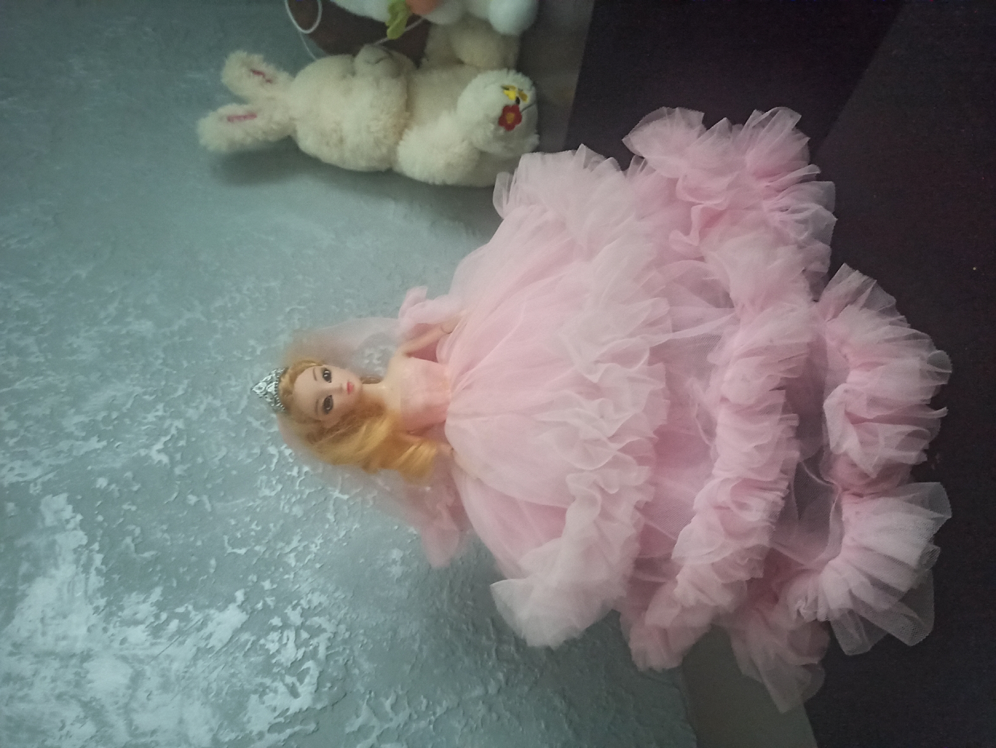 A doll with blonde hair and a tiara is dressed in a voluminous pink ruffled gown. It is placed on a dark surface in front of a textured gray wall. To the right of the doll are two stuffed animals: a cream-colored bunny and a white snowman with a carrot nose. The image is a close-up shot, likely taken indoors, and there is no discernible information about its location in Kryvyi Rih, Ukraine.