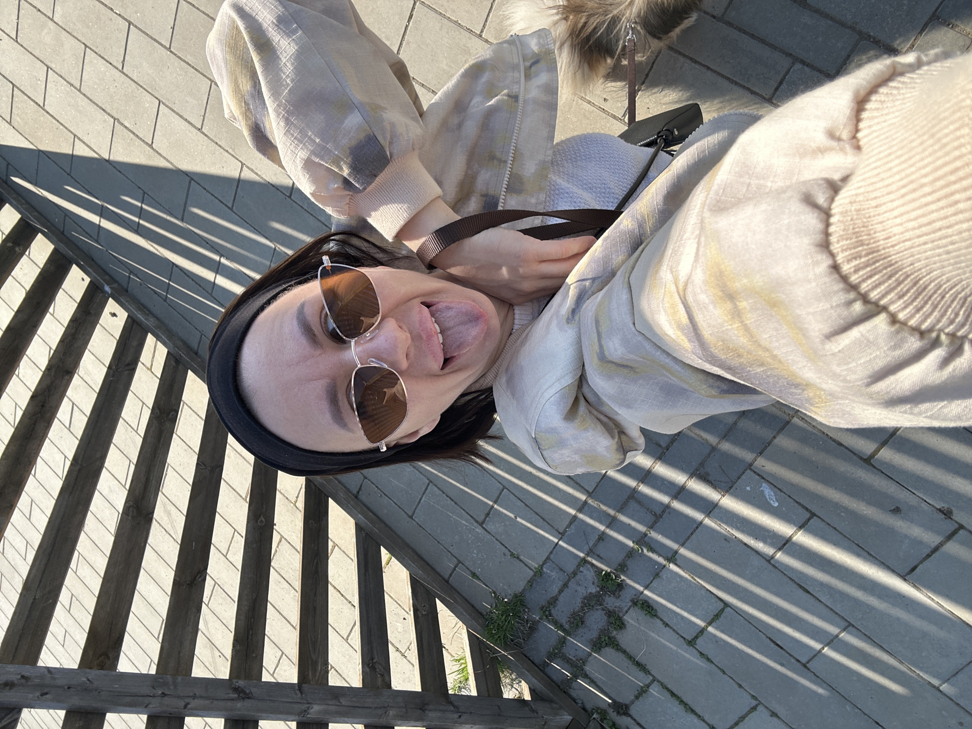 A high-angle, selfie-style photograph shows a woman outdoors. She has dark hair, sunglasses, and a black headband. Her tongue is sticking out in a playful manner. She is wearing a patterned jacket. A dog on a leash is partially visible in the upper part of the frame. The background features a brick walkway partially shaded by a wooden structure. Small weeds grow from the walkway's edges. The location is Votkinsk, Russia.