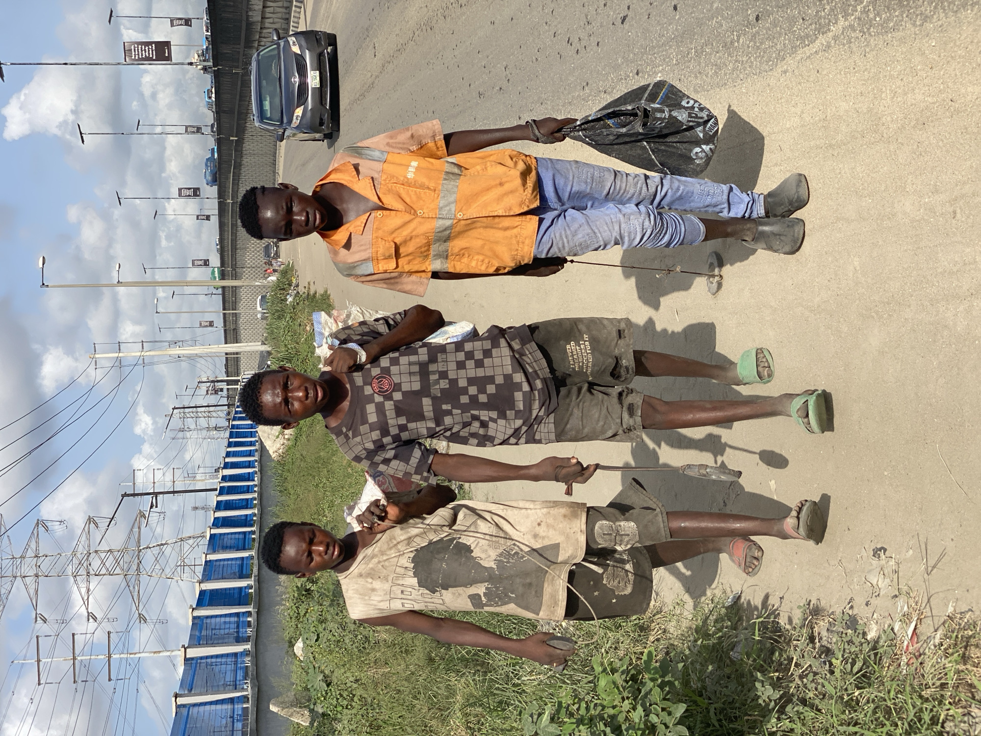 The image depicts three young men of African descent standing on the side of a road in Lekki, Nigeria. The topmost man wears a bright orange safety vest over a tan shirt and blue jeans, with black work boots. He carries a black bag in one hand and a metal wire or cable attached to an unseen object in the other. The second man is dressed in a dark checkered shirt and dark shorts, with green sandals. He holds a similar wire or cable. The third man wears a faded white t-shirt with a graphic and dark shorts, with one red sandal and one unseen. He too has a metal wire or cable.
They are standing on a stretch of pavement beside a multi-lane road with a gray vehicle in the background. A highway overpass is visible in the distance with light poles and billboards. A large, fenced structure sits in the background, with metal power pylons extending into the sky. The sky is partly cloudy.