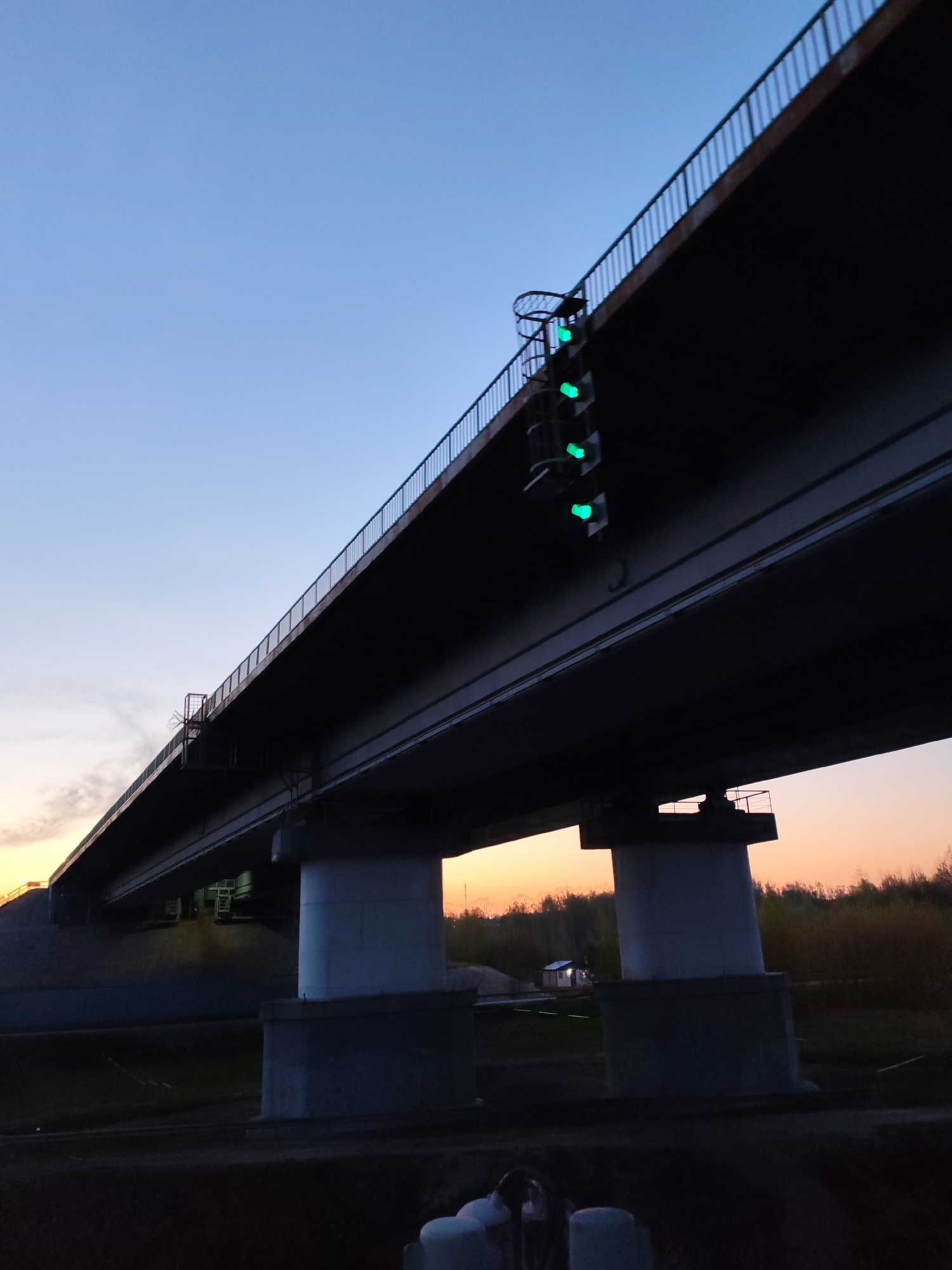 A low-angle shot depicts a concrete overpass against a gradient sky, transitioning from blue to orange. The overpass is supported by massive concrete pillars, with some foliage visible in the background. Attached to the side of the overpass is a vertical structure containing three green lights. The foreground is obscured in shadow.