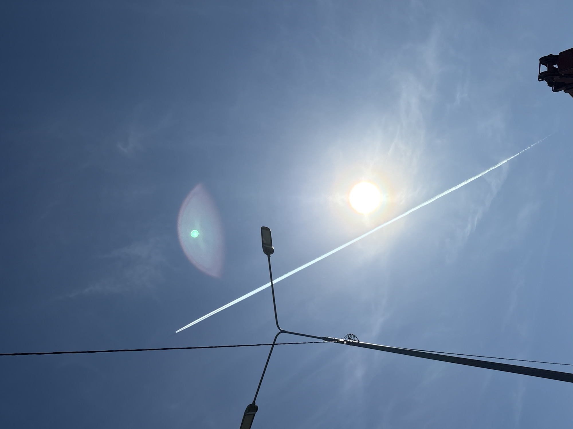 The image shows a bright, sunny day in Verkh-Sosnova, Russia. The sky is a clear blue, with a vapor trail from a jet airplane crossing the frame diagonally. The sun is visible, causing a lens flare effect. In the foreground is a lamppost with two lamps extending at different angles. Electrical wires are also present, crossing the lower part of the image. The upper right corner shows part of some metallic machinery. There are no people visible.