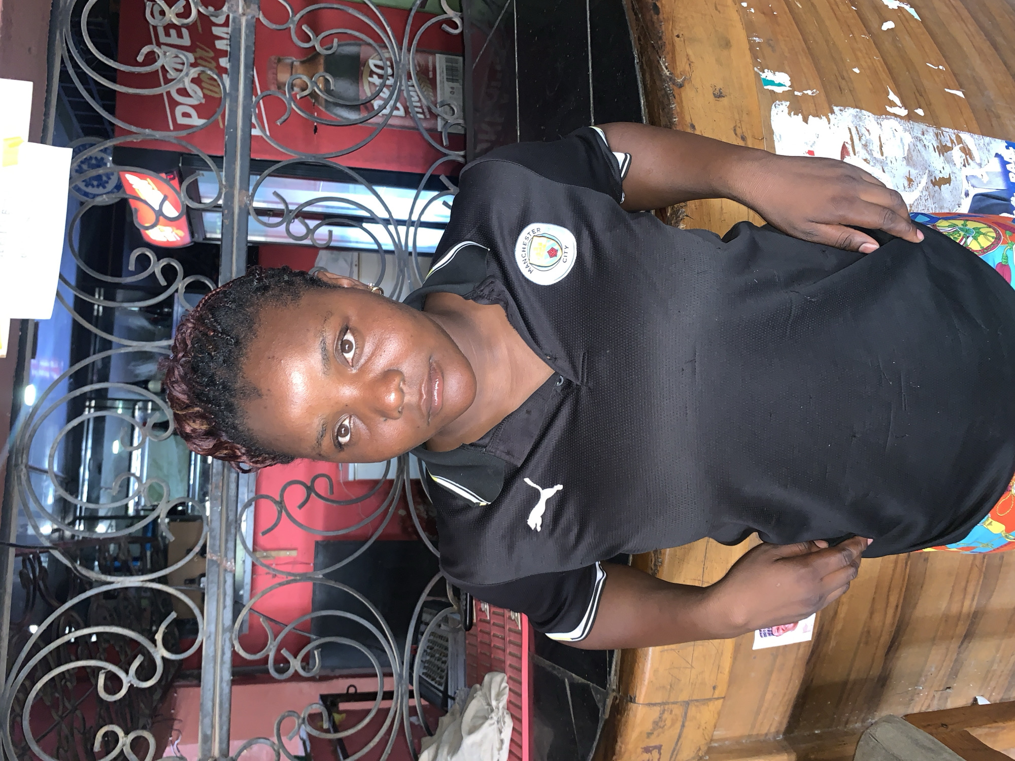 The image depicts a woman standing in Jos, Nigeria. She wears a black Puma polo shirt with a Manchester City emblem. She is standing in front of a black metal security gate with decorative swirls. Behind the gate, a sign for "Power & Co." is visible. The background shows signs of a commercial area, possibly a shop or restaurant. The woman's hand is resting on her hip. She is looking directly at the camera. The floor appears to be wooden and worn with patches of missing paint or paper. The environment suggests an urban setting in Nigeria.