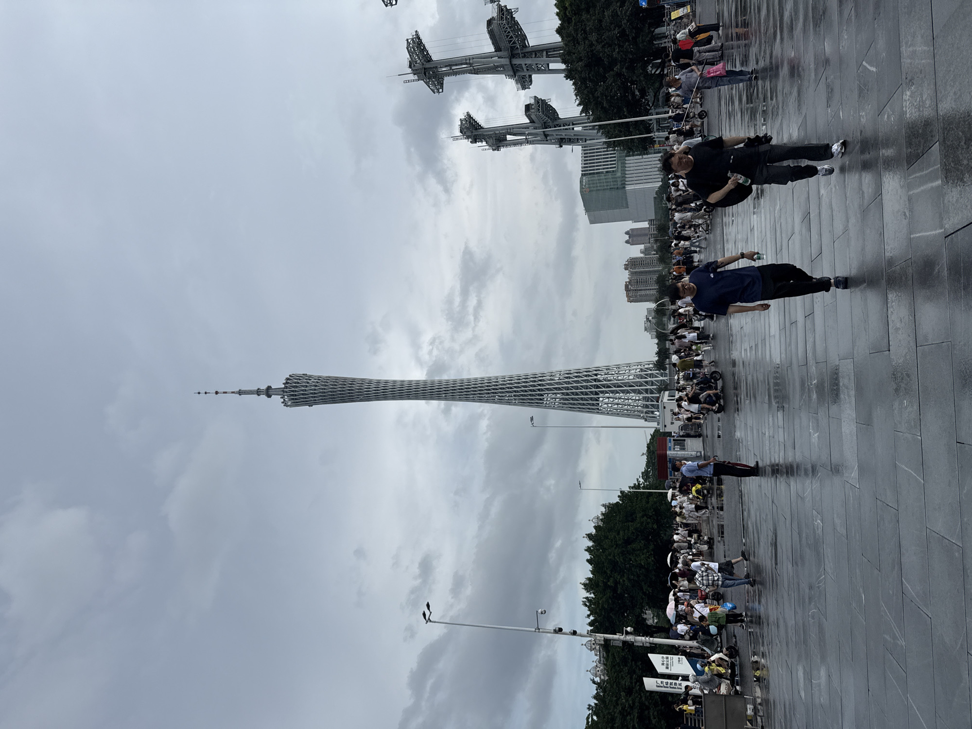 The image shows a wide, paved plaza on a cloudy day, with wet surfaces reflecting the overcast sky. A tall, lattice-like tower, identified as the Canton Tower, dominates the background. To the right of the tower, there are tall, metallic structures that appear to be part of an entertainment venue or observation area. In the middle ground, a crowd of people is visible, some walking, others on scooters or pushing strollers. Many people are holding umbrellas, suggesting it may be raining or has recently rained. Trees line the edge of the plaza behind the crowd. The foreground shows two men walking towards the viewer on the wet pavement. One man is wearing a dark blue t-shirt and black pants, carrying a water bottle. The other man is wearing a black t-shirt and dark pants, carrying a black bag over his shoulder and a water bottle in his hand. The scene is set in Tianhe District, China.