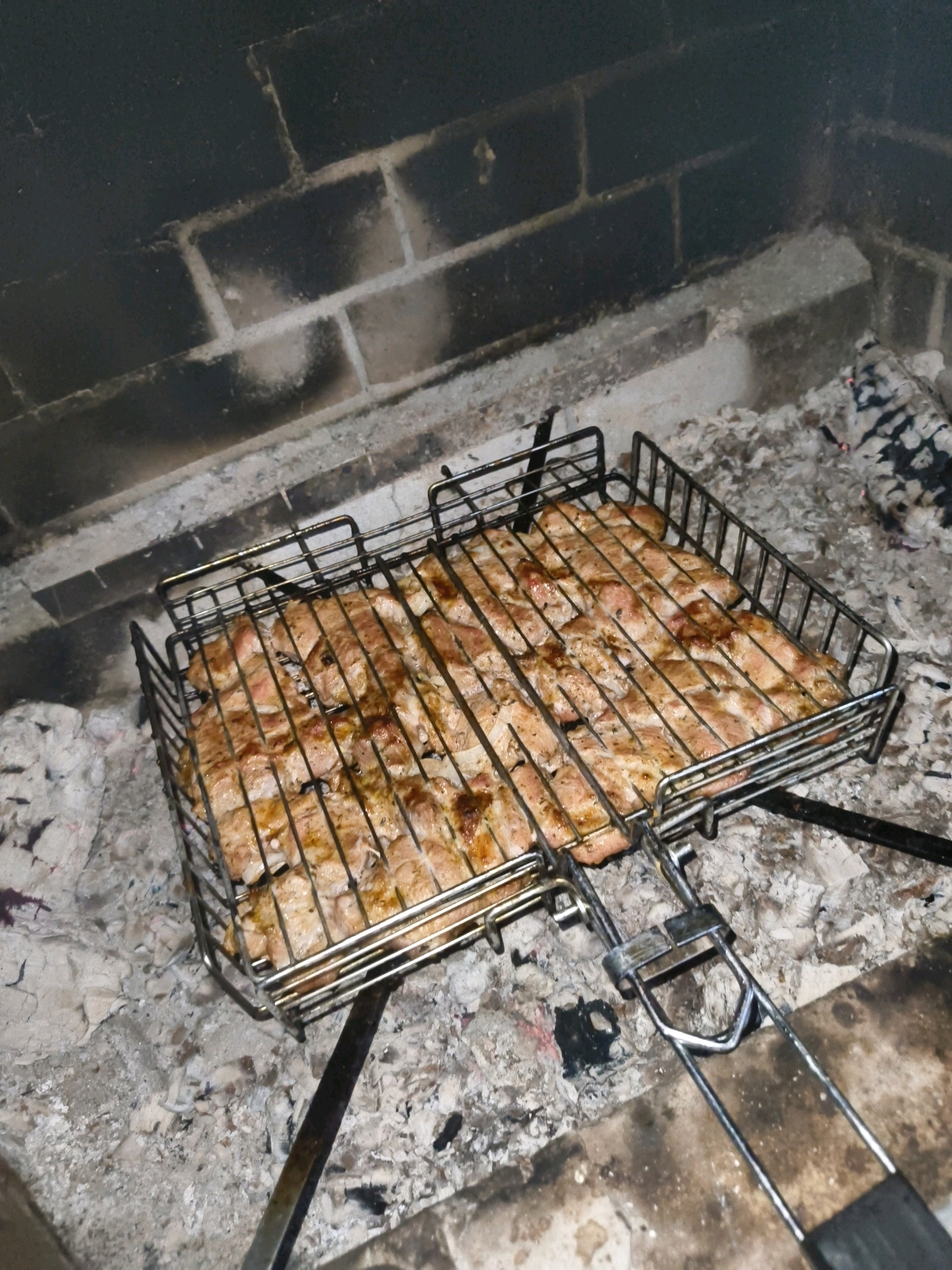 A grilling basket filled with raw meat is placed over glowing embers and ash within a brick fireplace. The meat is thinly sliced and arranged in rows on the metal grate. The fireplace walls are constructed of dark bricks, with some soot and ash visible on the surfaces. The overall scene suggests an outdoor cooking setting, possibly in the city of Vostets SNT, Russia, given the context of the image.