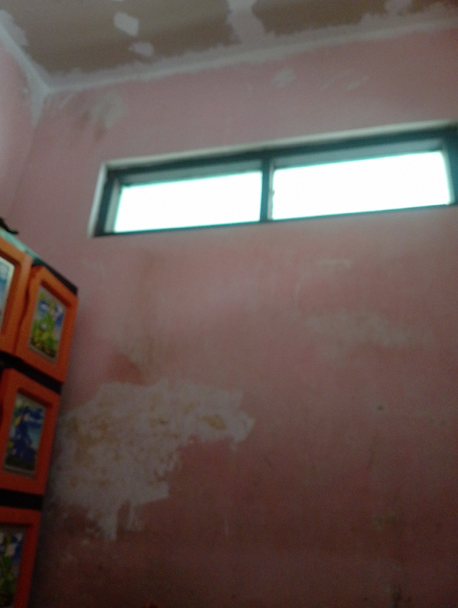 The image shows the interior of a room with pink walls and a ceiling that appears to be undergoing renovation or repair. There are patches of white plaster visible on the ceiling and along the top of the walls. A horizontal window with white panes is set high on the wall. To the left of the image is a multi-colored plastic storage unit with orange frames and visible picture inserts on its drawers. The wall surface exhibits some discoloration or unevenness. The lighting is somewhat dim, creating a grainy appearance. The scene does not provide any specific clues to its precise location within Sukamanah, Indonesia, beyond being an indoor residential or utility space. No people or actions are depicted.